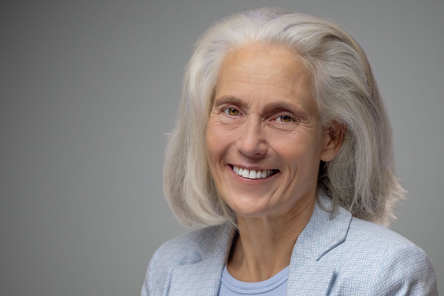 Smiling older woman with gray hair wearing a light blue blazer against a gray background.