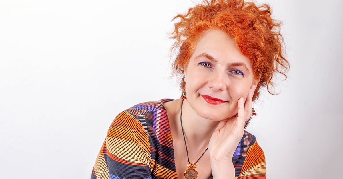 Portrait of someone in a colorful striped sweater with vibrant curly red hair against a white background.