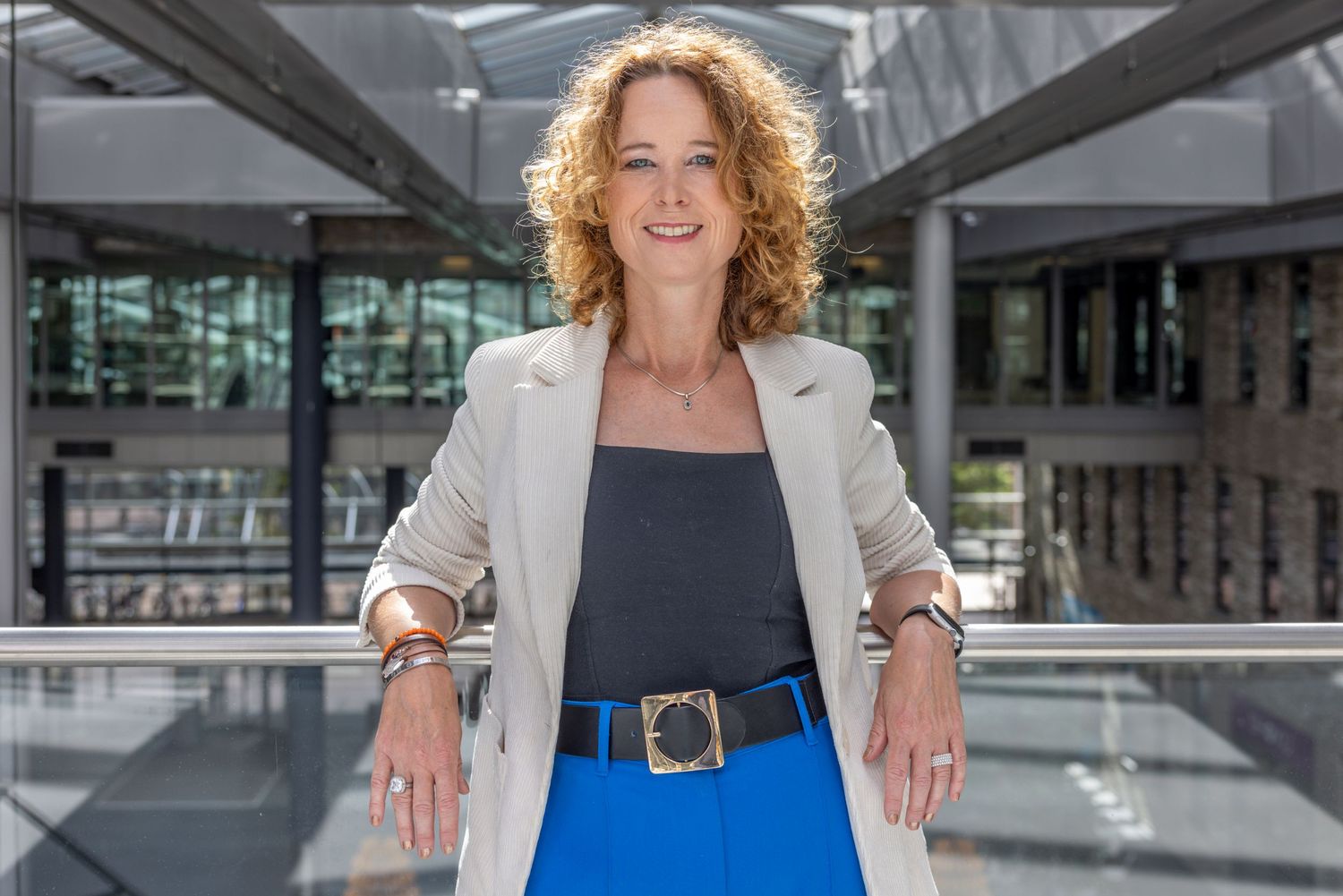 Professional woman in business attire stands confidently in modern office setting with glass walls and natural light.