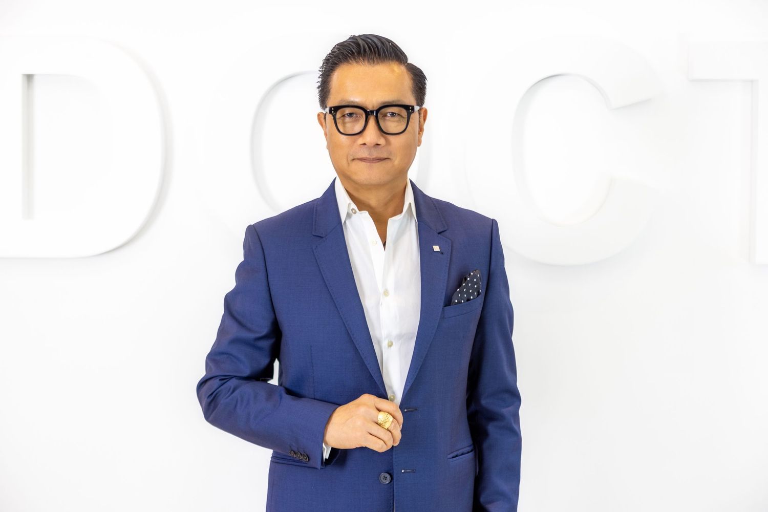 A person wearing a navy blue suit jacket and glasses poses professionally against a white background.
