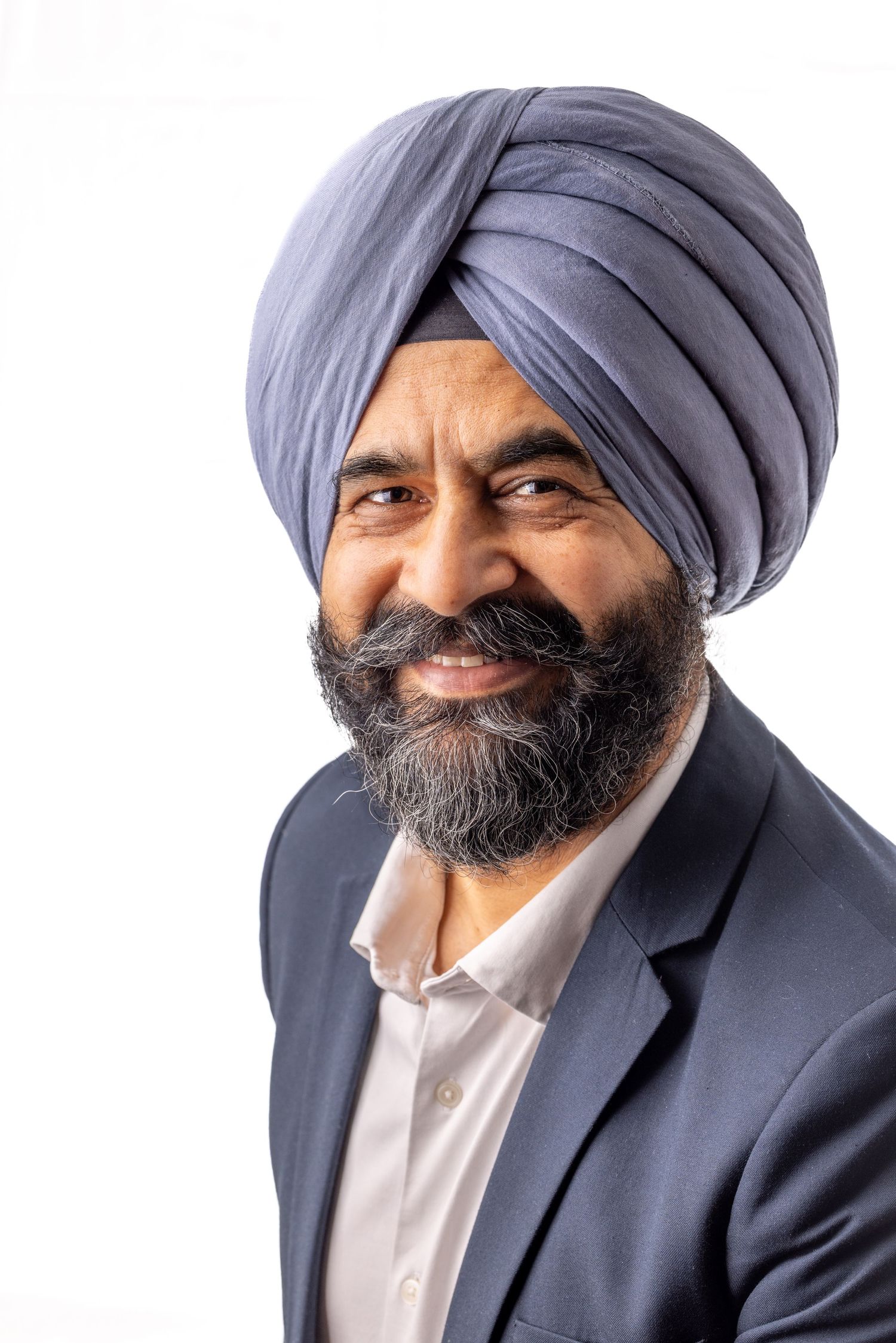 Smiling Sikh man with beard wearing blue turban and suit jacket against white background.