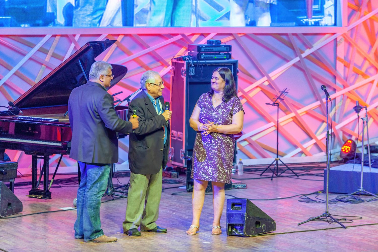 Musicians interact on a colorfully lit stage with a piano and performance equipment visible.