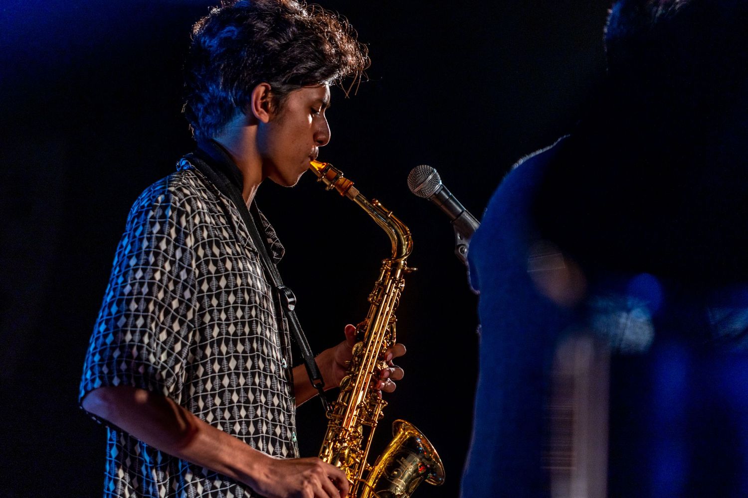 Musician playing saxophone in patterned shirt under stage lighting.
