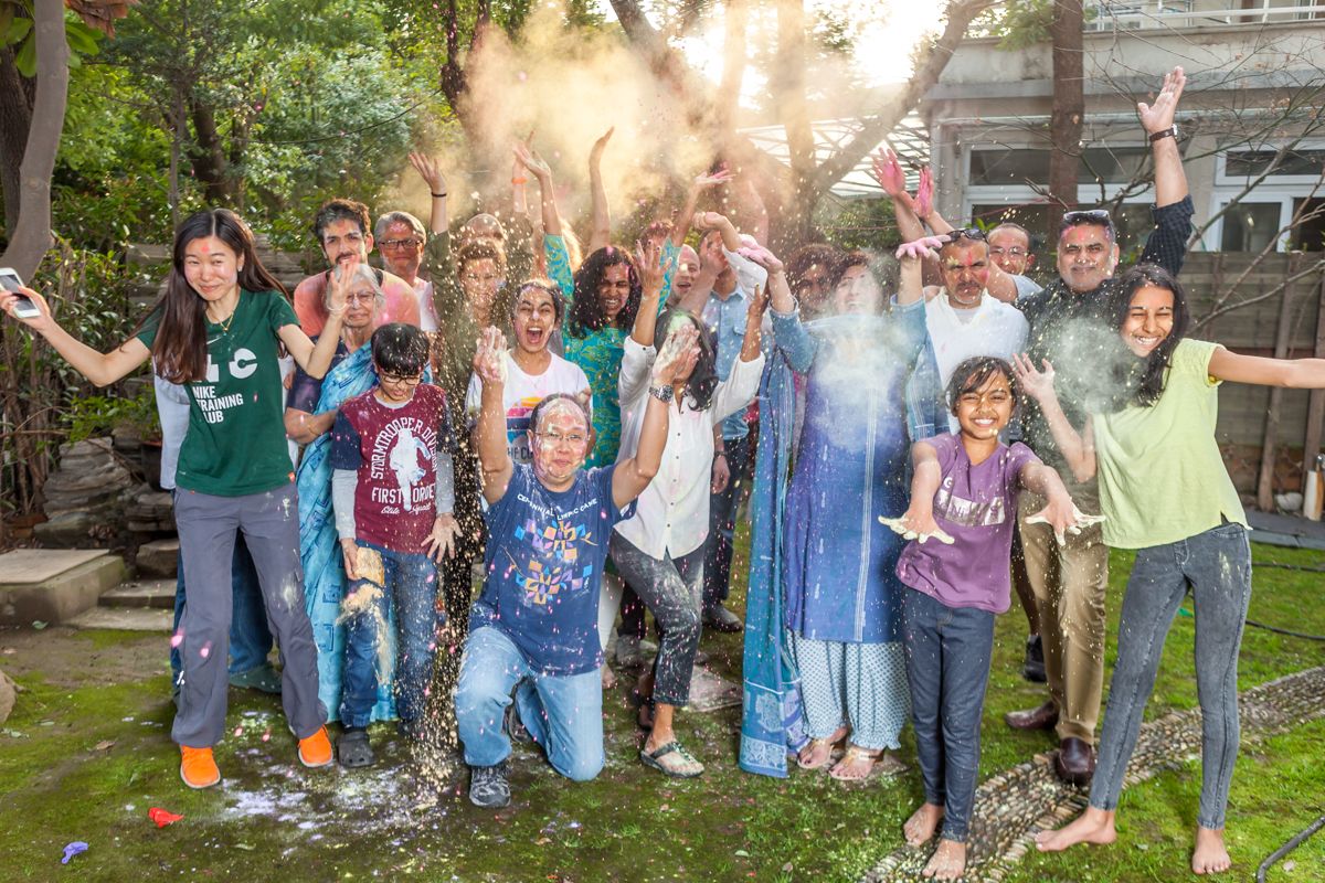 A cheerful group poses outdoors in casual clothing with their hands raised against a sunlit garden background.