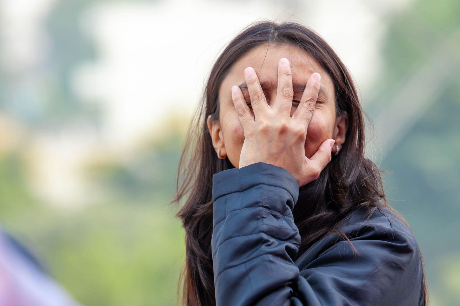 A person laughing and covering their face while wearing a dark jacket outdoors.