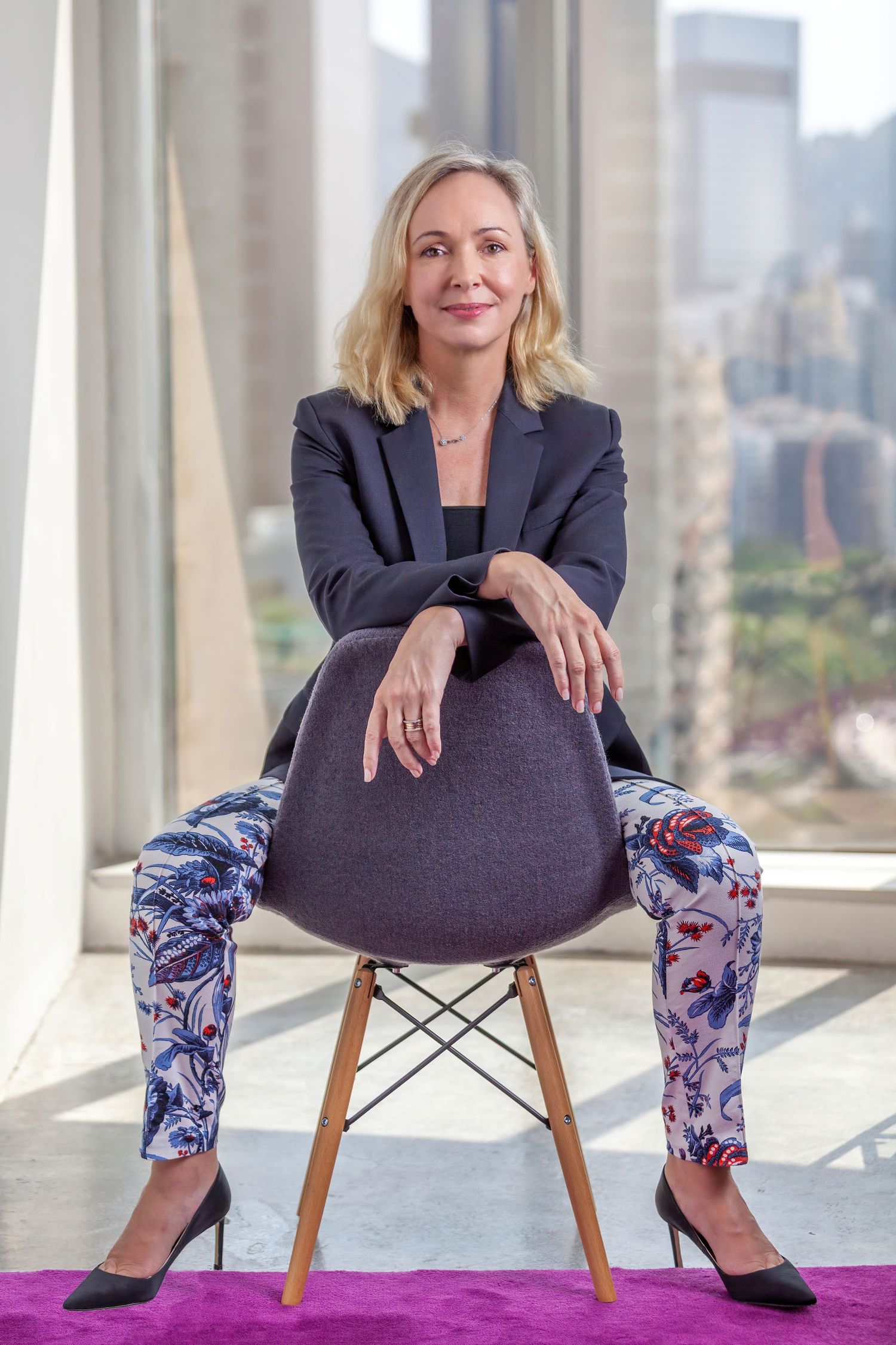 Professional sitting on modern chair in business attire with floral print pants in sunlit office setting.
