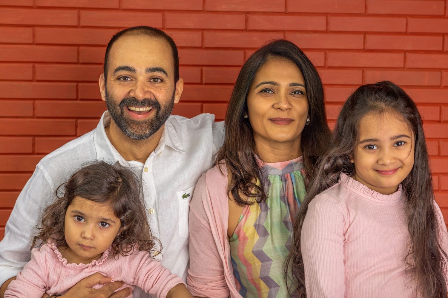 A group of people pose together in front of a red brick wall wearing casual attire in pink and gray tones.