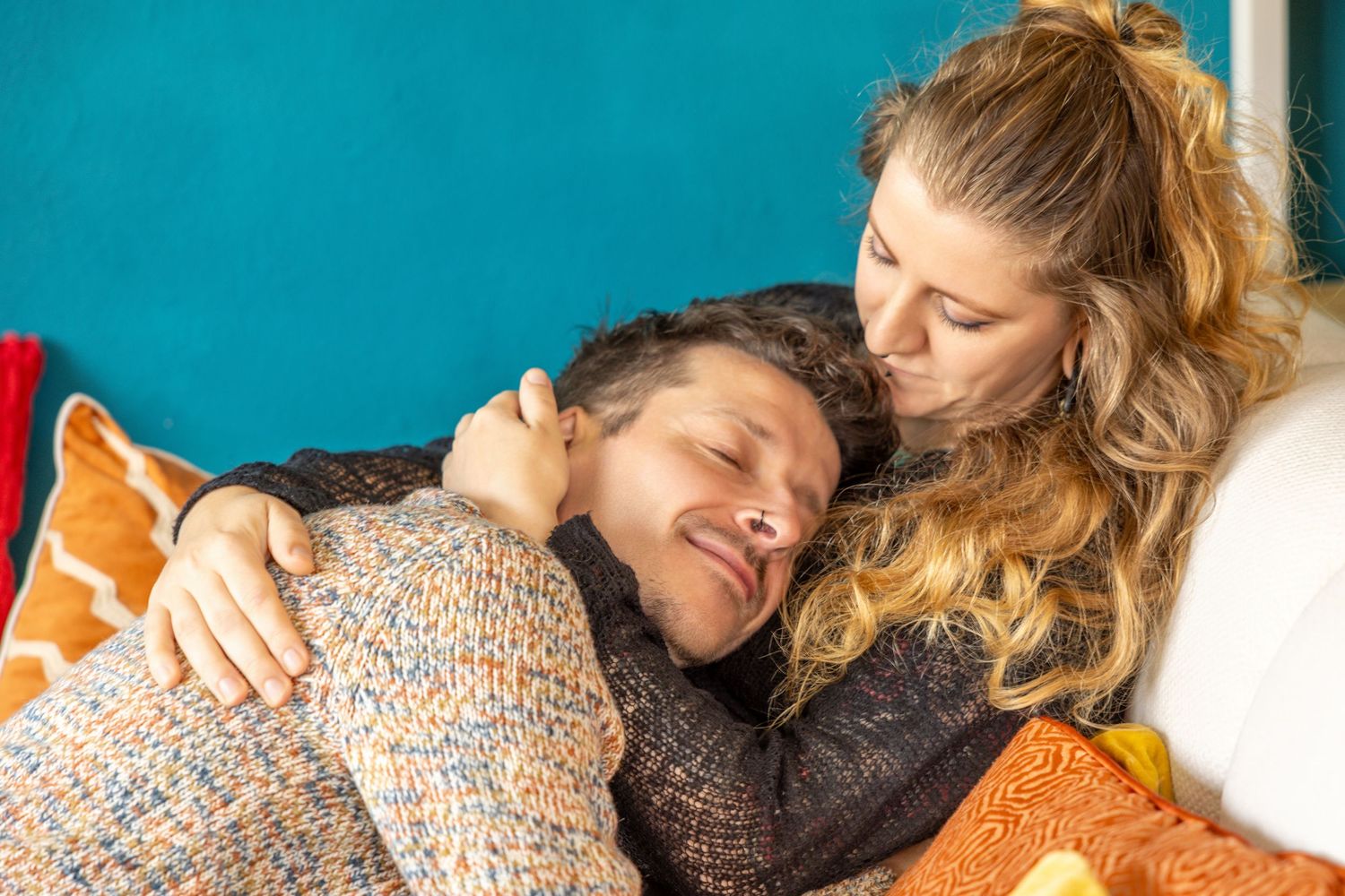 A happy couple cuddling on a couch, woman embracing man from behind, smiling warmly in cozy sweaters.