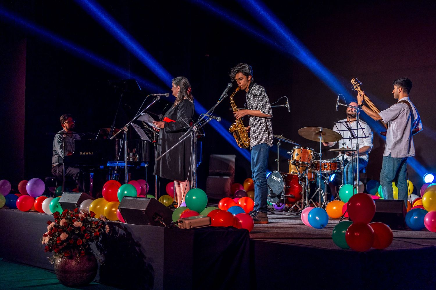 Band performs on stage with colorful balloons and blue lighting creating a festive atmosphere.