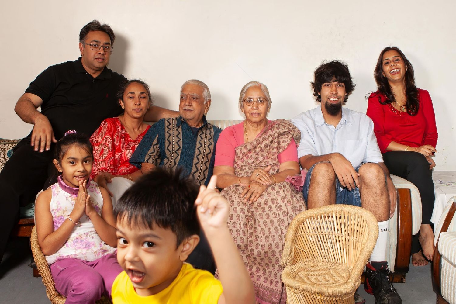 Multi-generational family group sitting together on a couch with a woven rattan chair in a casual home setting.