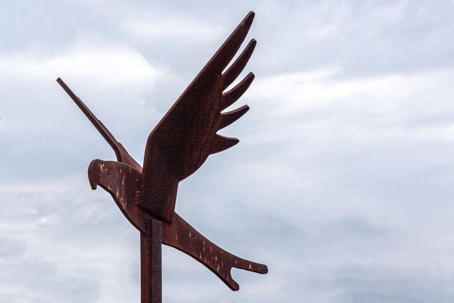 Metal weathervane in the shape of a bird with spread wings against cloudy sky.