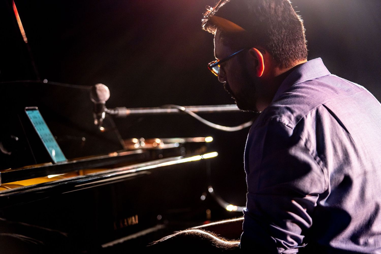 Side view of pianist performing at grand piano with stage lighting in background.