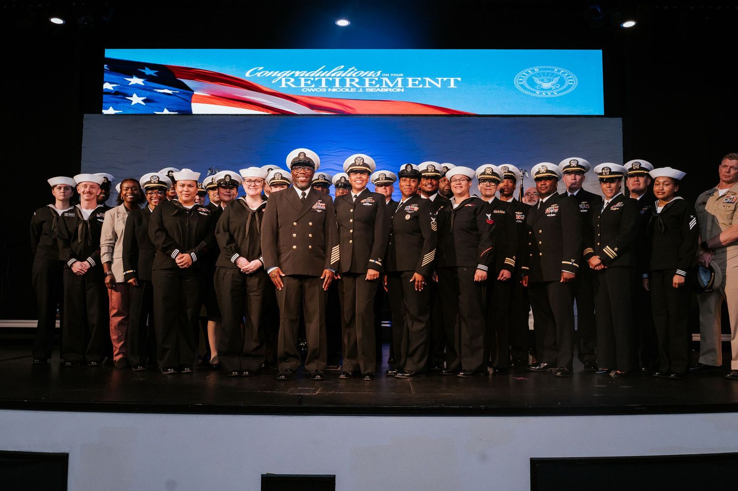 Group of uniformed Navy officers and sailors standing together on a stage in front of an American flag banner at a retirement ceremony.