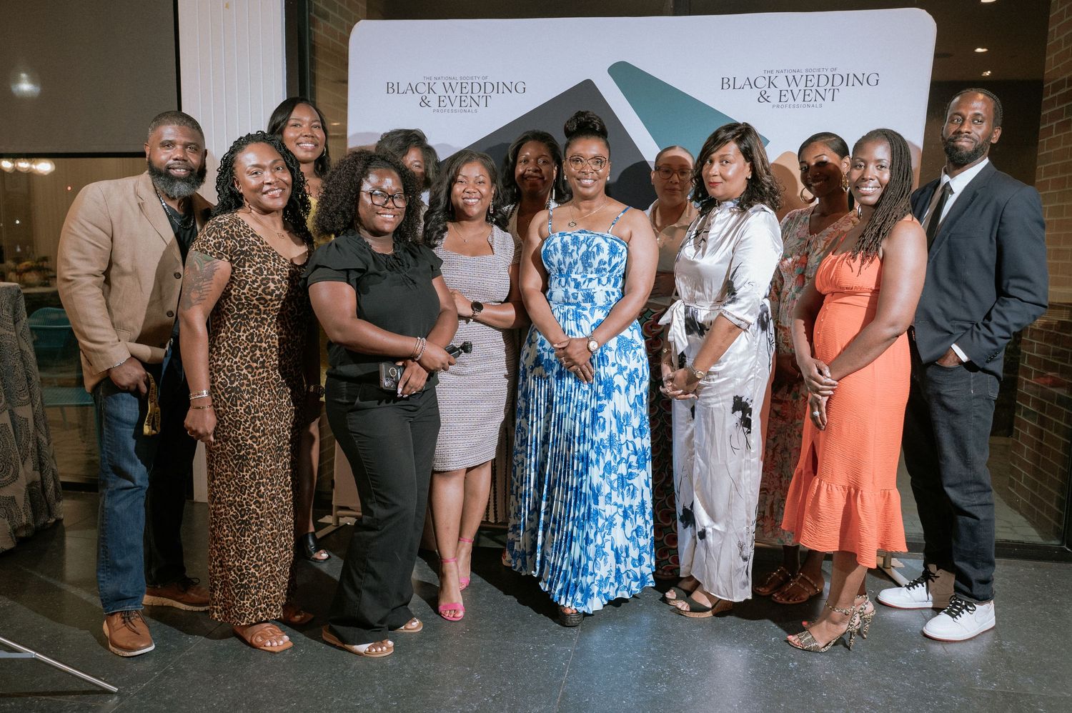 Group of smiling men and women in stylish formal attire posing together in front of a Black Wedding & Event banner at an indoor event.