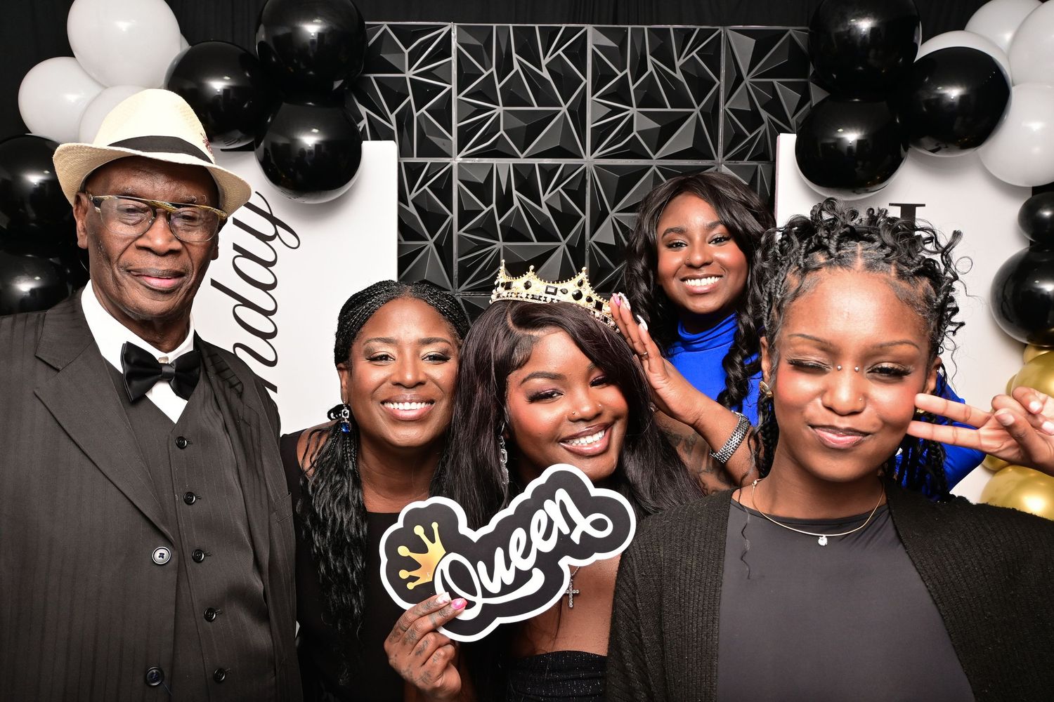 Group of five people posing at a birthday party with black and white balloons and a Queen sign.
