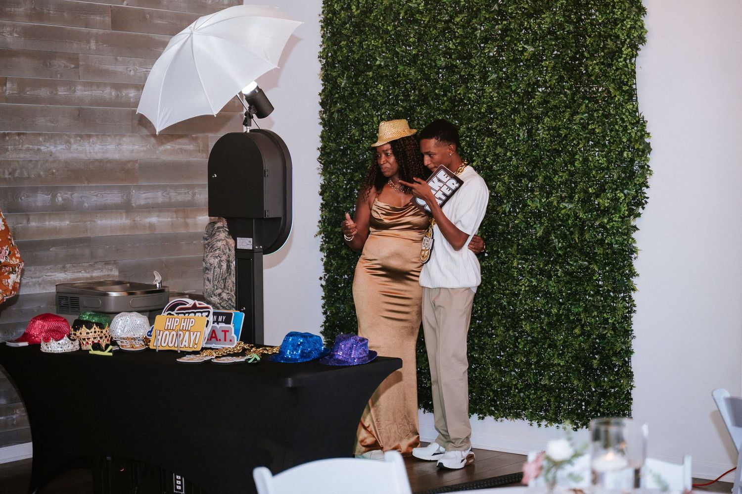 Couple embracing in front of a green hedge wall at an elegant indoor event with photo booth props.