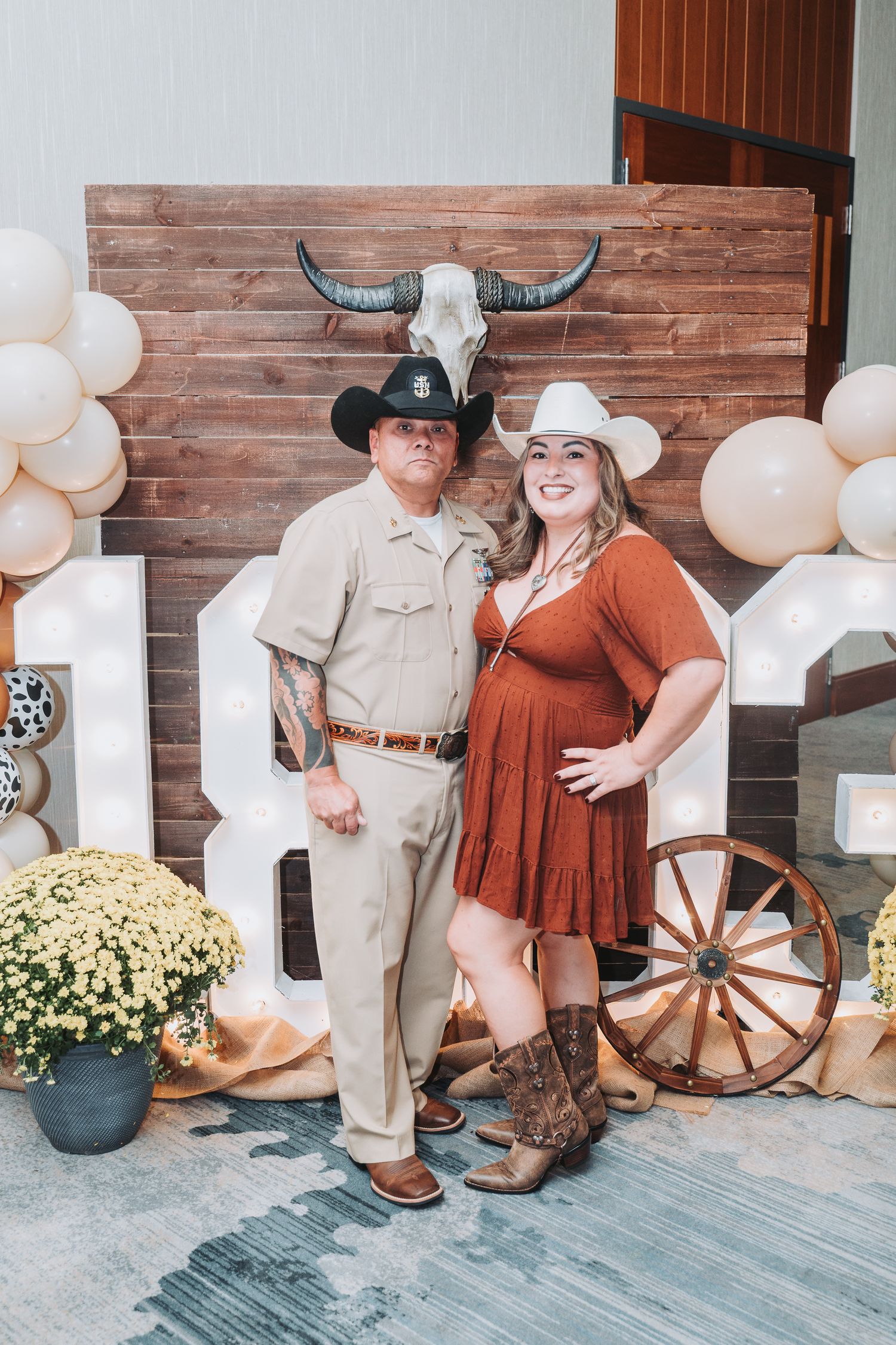 A couple in western attire poses by a rustic backdrop with longhorn skull and wagon wheel decorations.