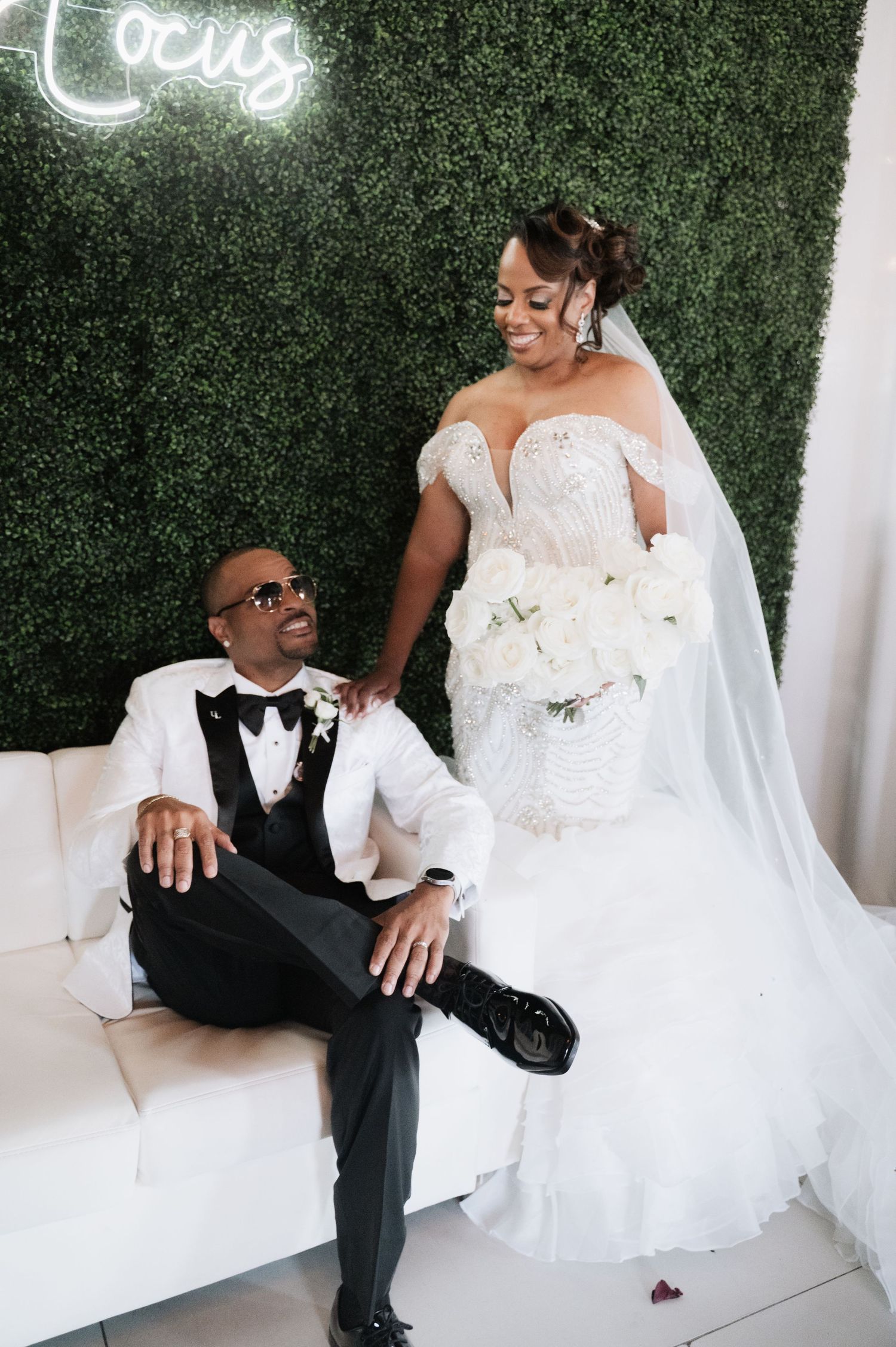 Wedding couple poses playfully against green hedge wall with neon signage.