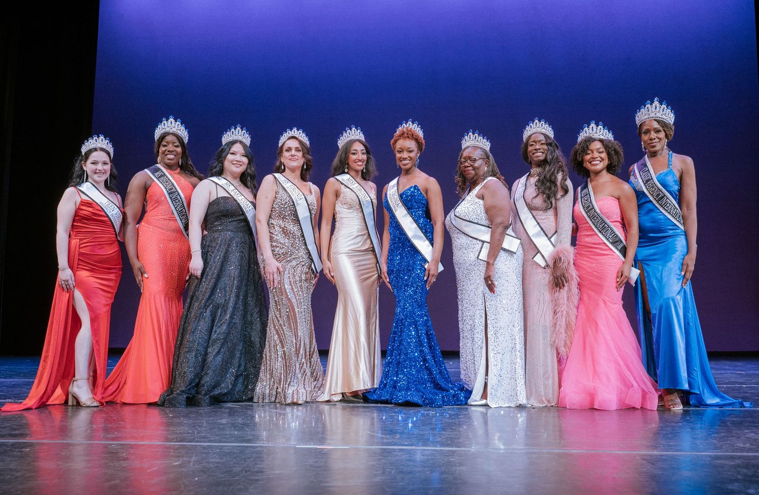 Ten women in formal gowns, crowns and sashes stand in a line on a stage under blue lighting at a beauty pageant.