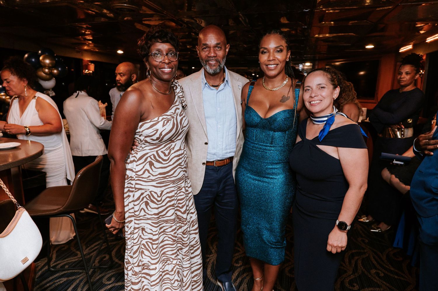 Four smiling guests in elegant evening attire pose together at a festive indoor event with other attendees seated at tables behind them.