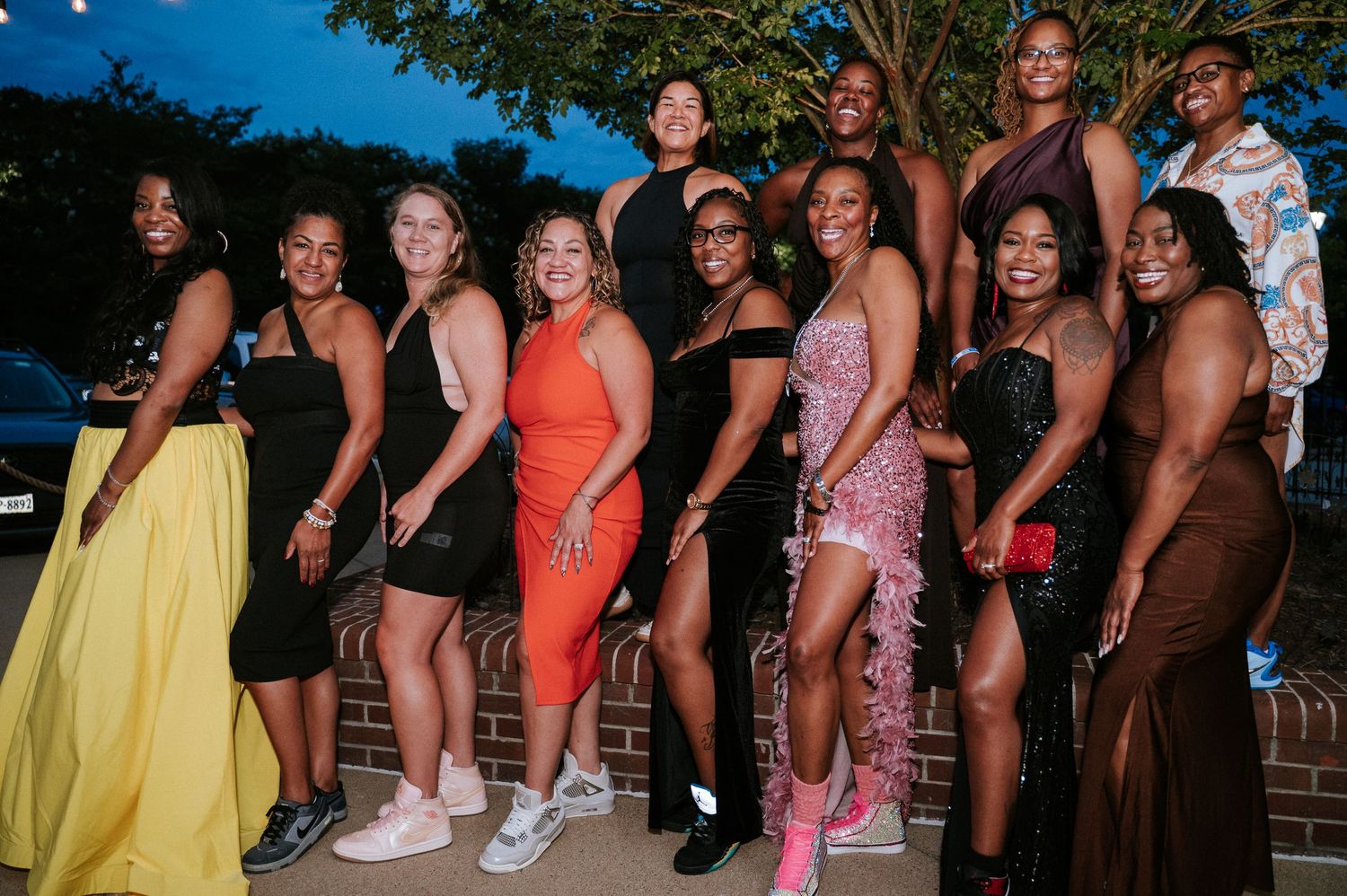 Group of smiling women in elegant dresses and heels posing together outdoors near a brick wall at dusk during a formal evening event.