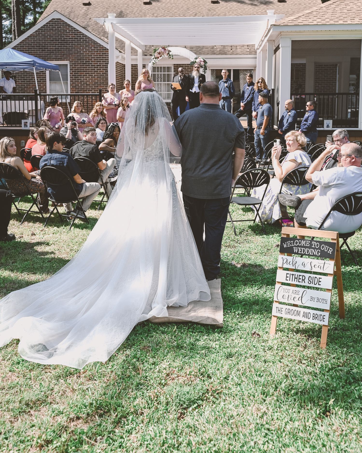 Outdoor backyard wedding ceremony with guests seated on folding chairs and welcome sign on display.