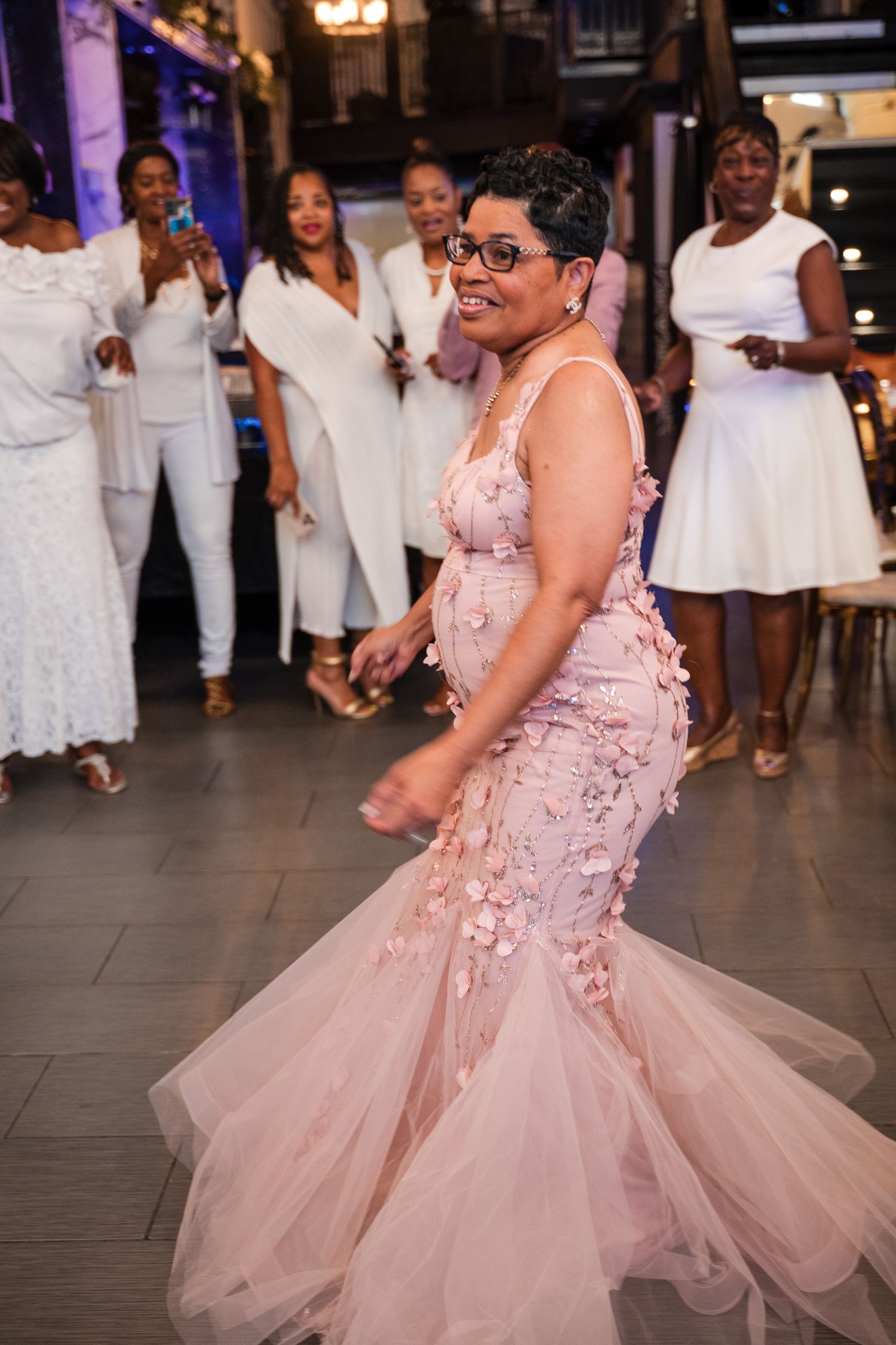 A person in a blush pink mermaid-style gown dances at a celebration while others in white outfits watch and cheer.