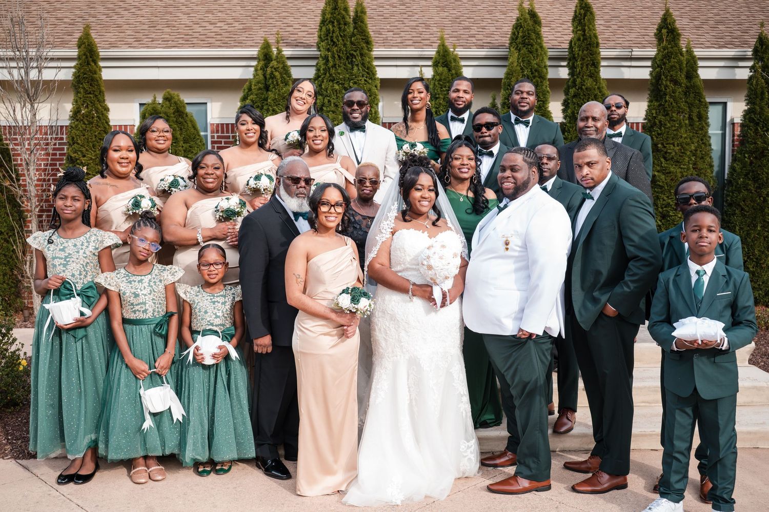 Large wedding party in emerald green dresses and dark suits poses outdoors around a bride in a white gown, with flower girls in front.