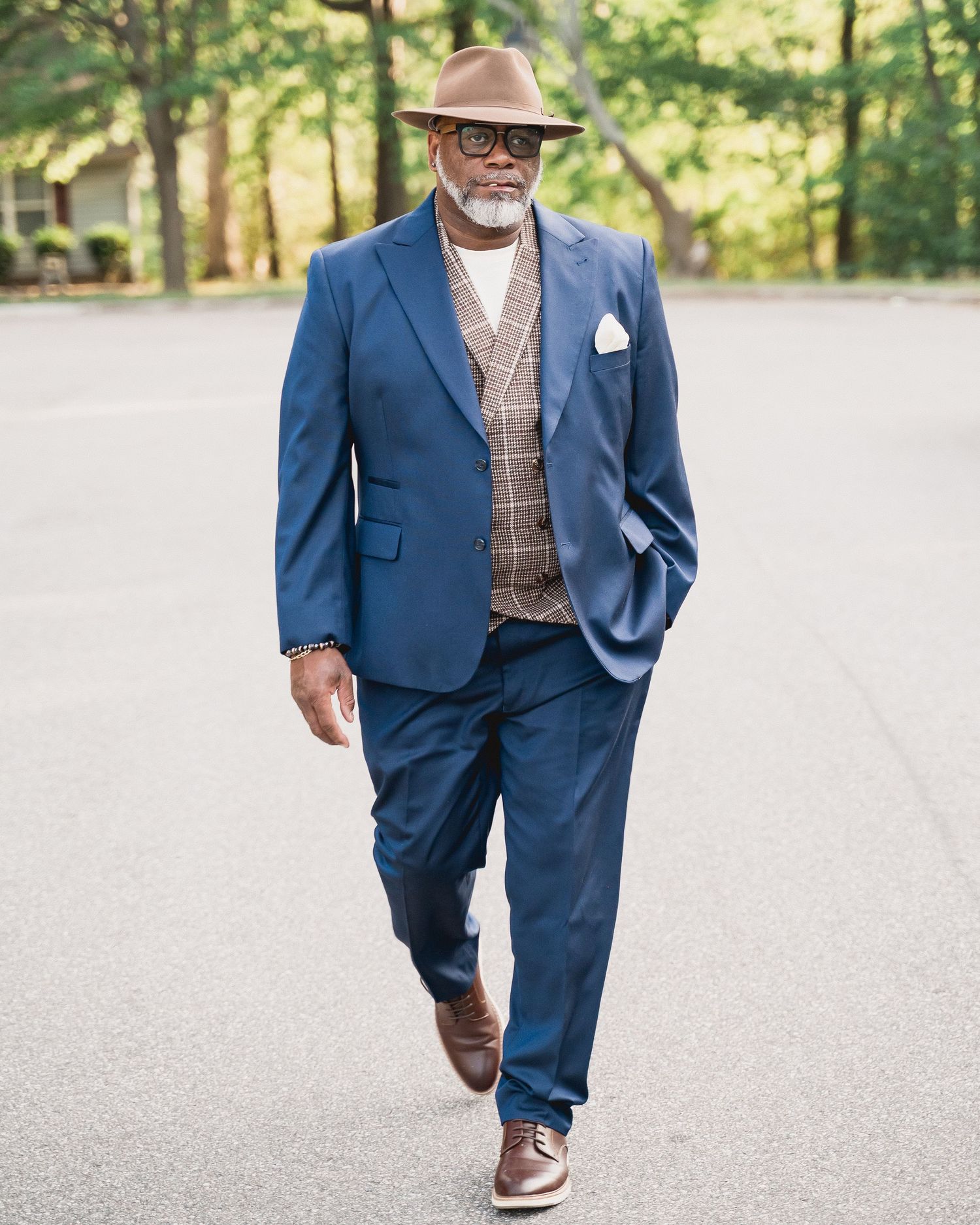 Stylish older man in blue suit, plaid vest, brown hat and shoes walks confidently on a tree-lined path.