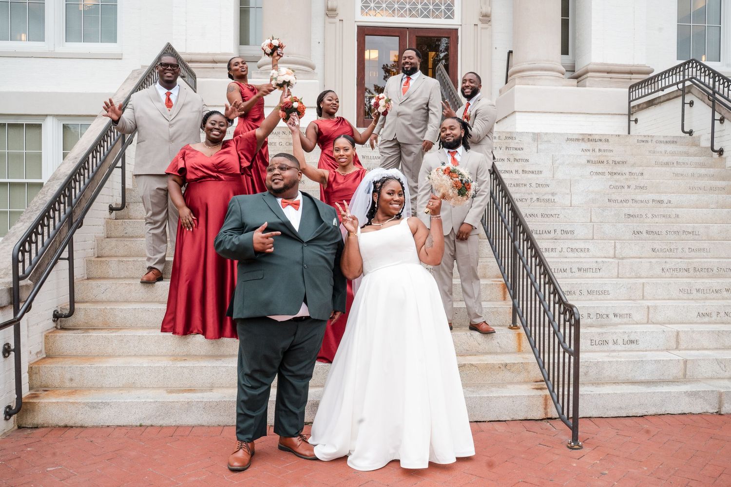 Wedding party members pose on elegant white stone steps of a building wearing formal attire.