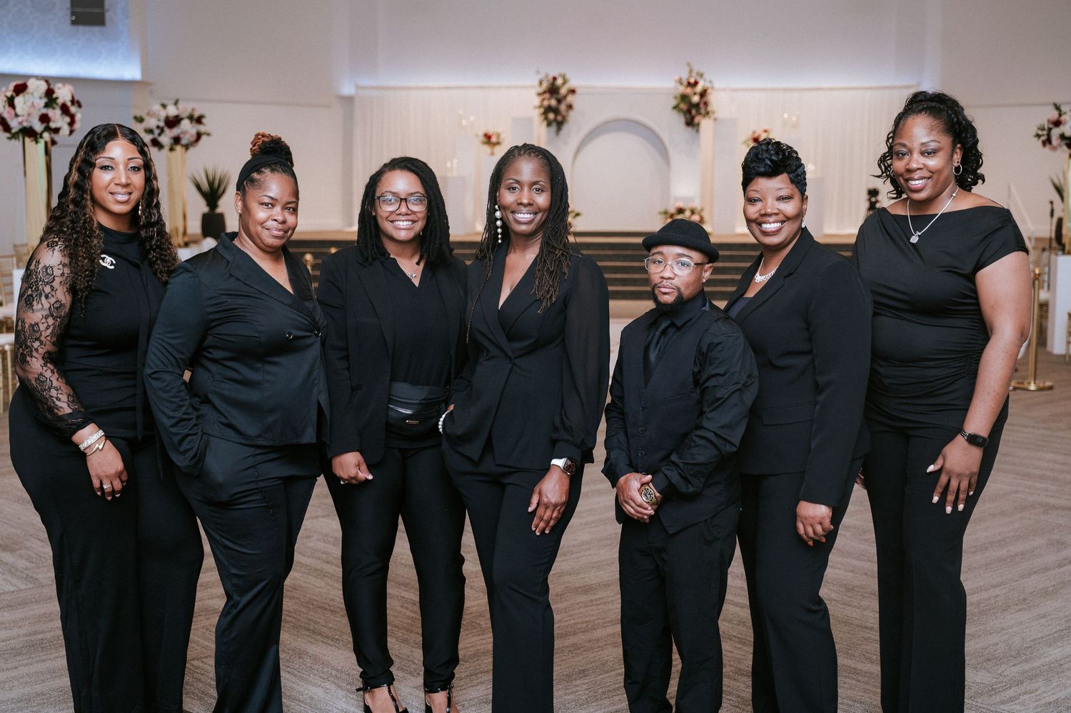Seven women in coordinated black outfits pose together in an elegant event venue decorated with floral centerpieces and white drapery.