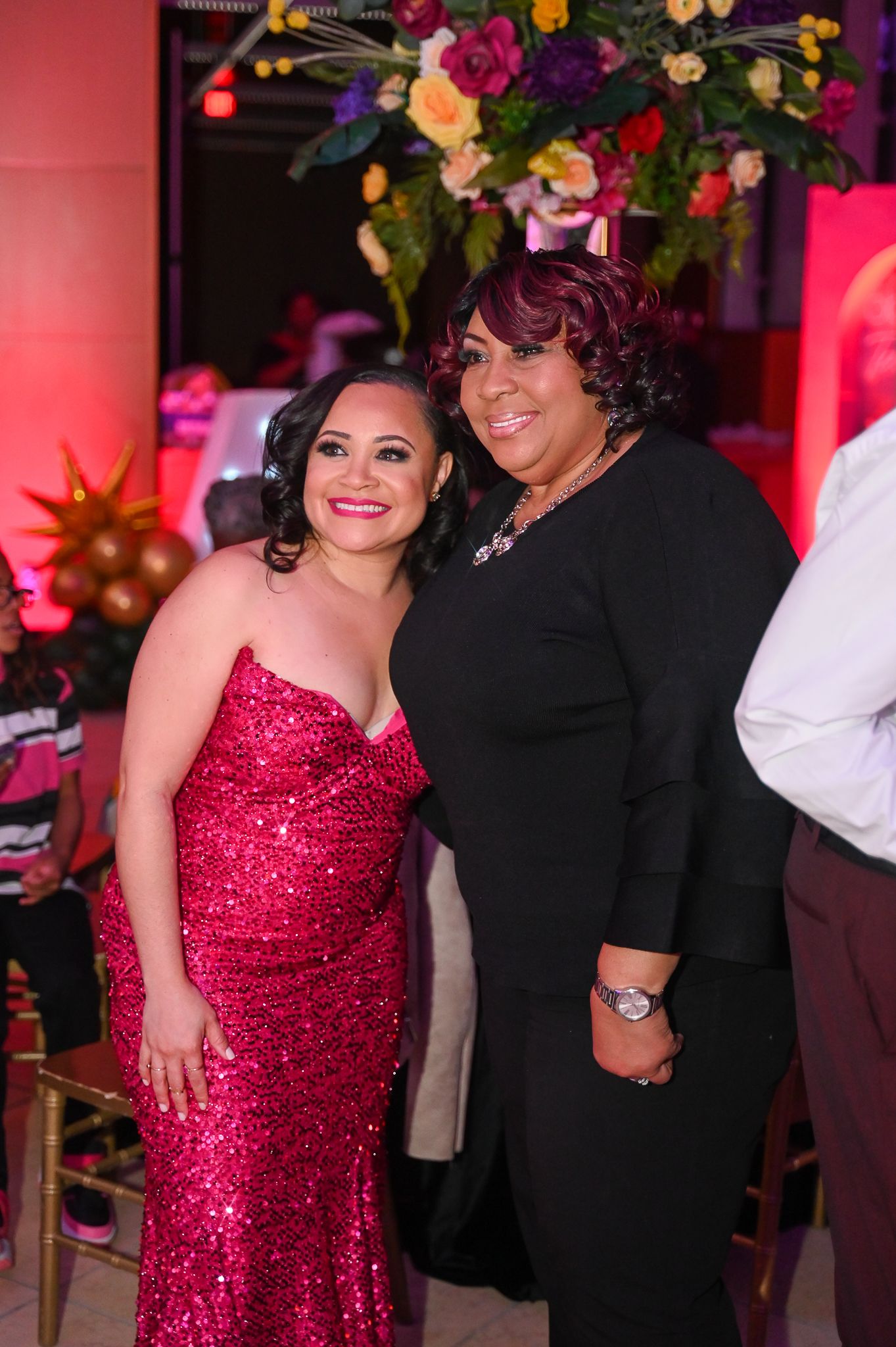 Two guests pose together at an elegant event with colorful floral decorations, one wearing a sparkly red gown.