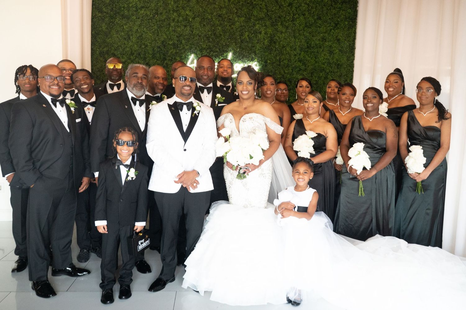 Large wedding party group photo with formal attire against a white and green backdrop.