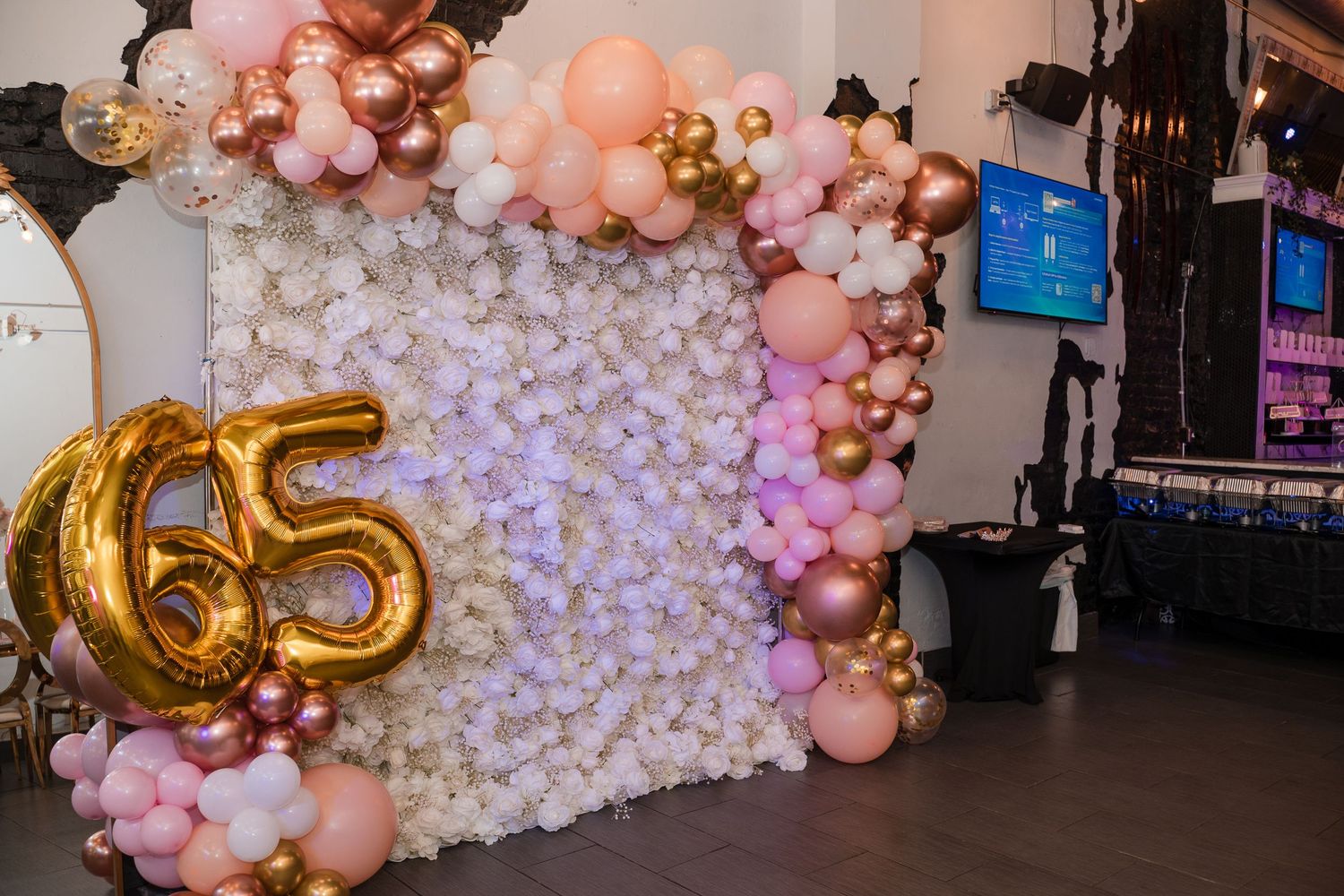Elegant balloon arch display with gold number 65, pink balloons and white floral backdrop for celebration.