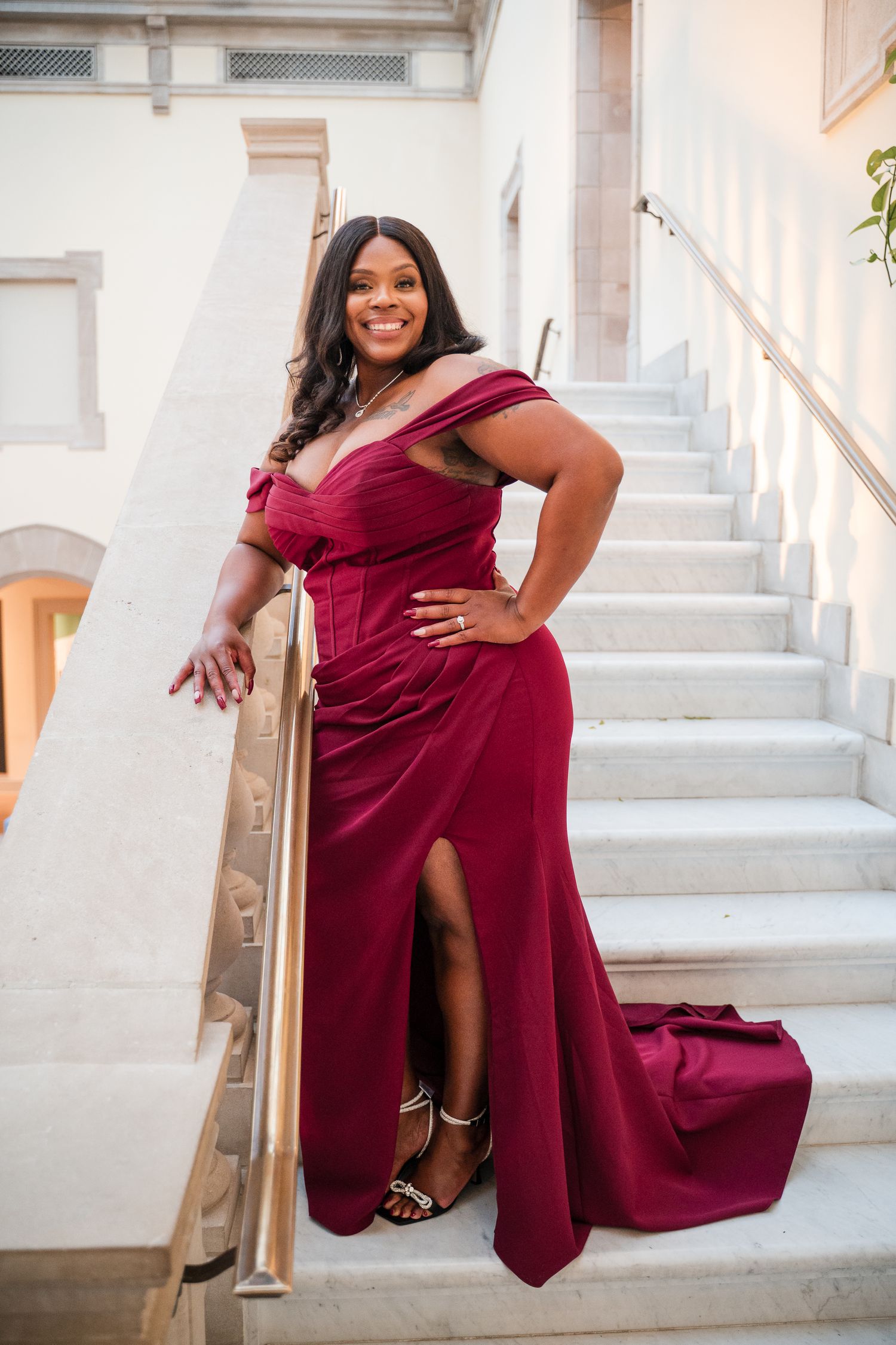 A woman in a burgundy gown poses confidently on white marble stairs during a formal photoshoot.