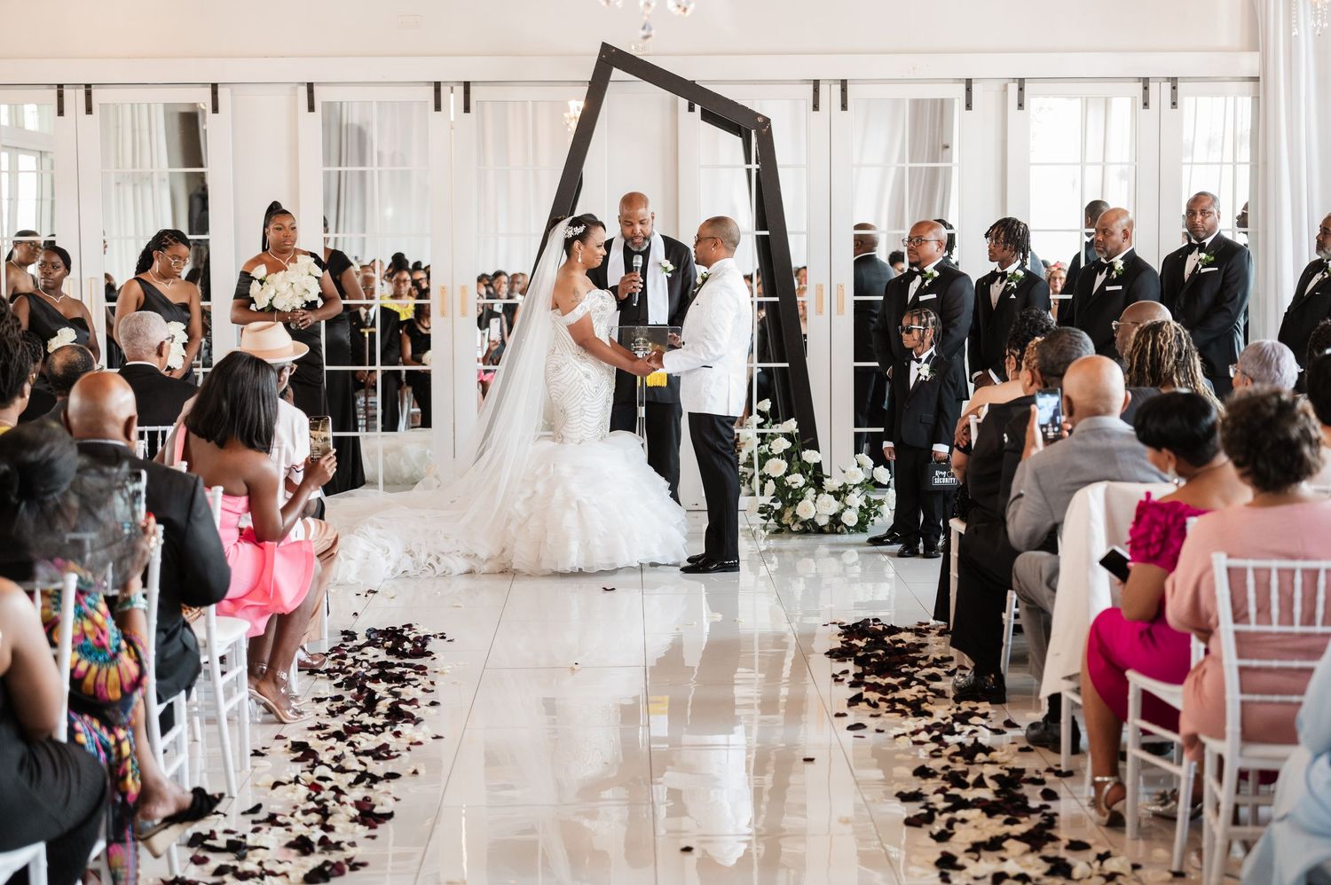 Wedding ceremony guests watch as rose petals line the aisle in an elegant white venue.