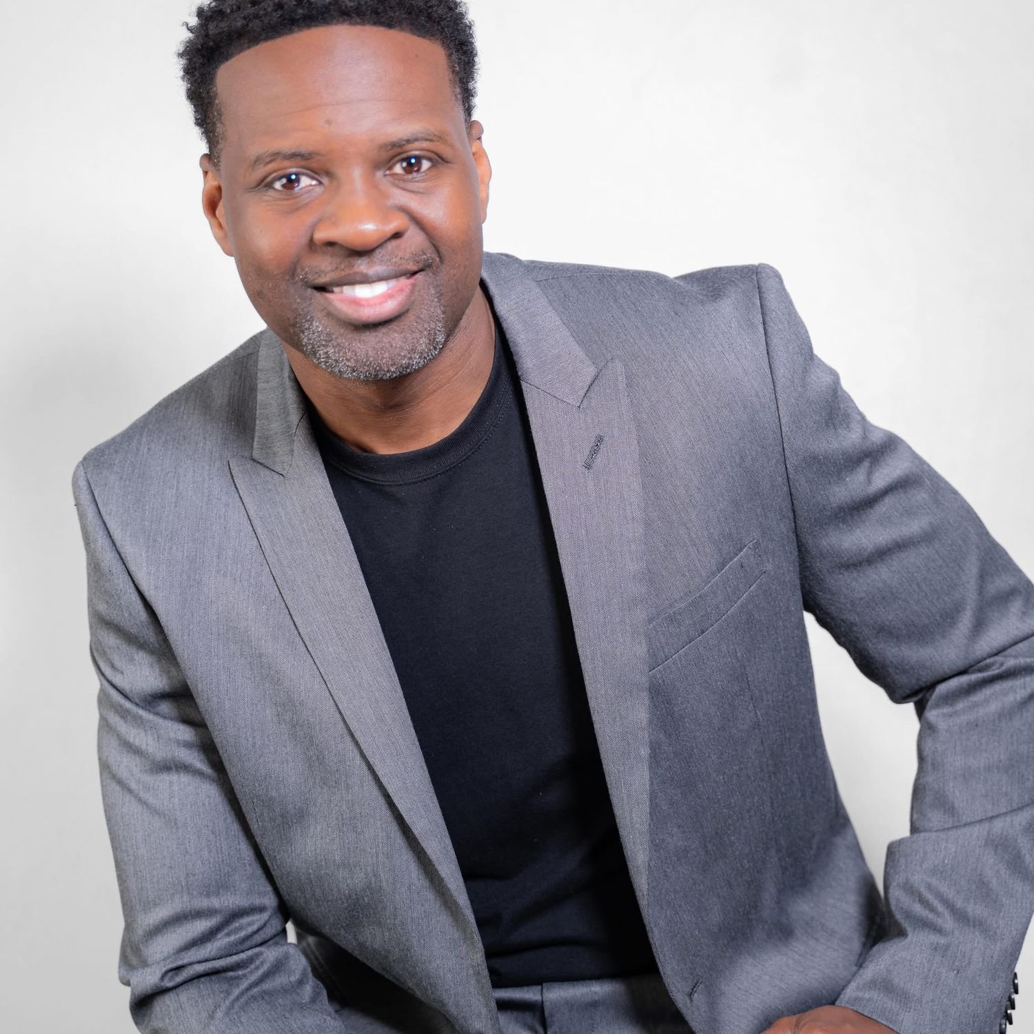 Professional headshot of a person wearing a gray blazer and black shirt against a white background.