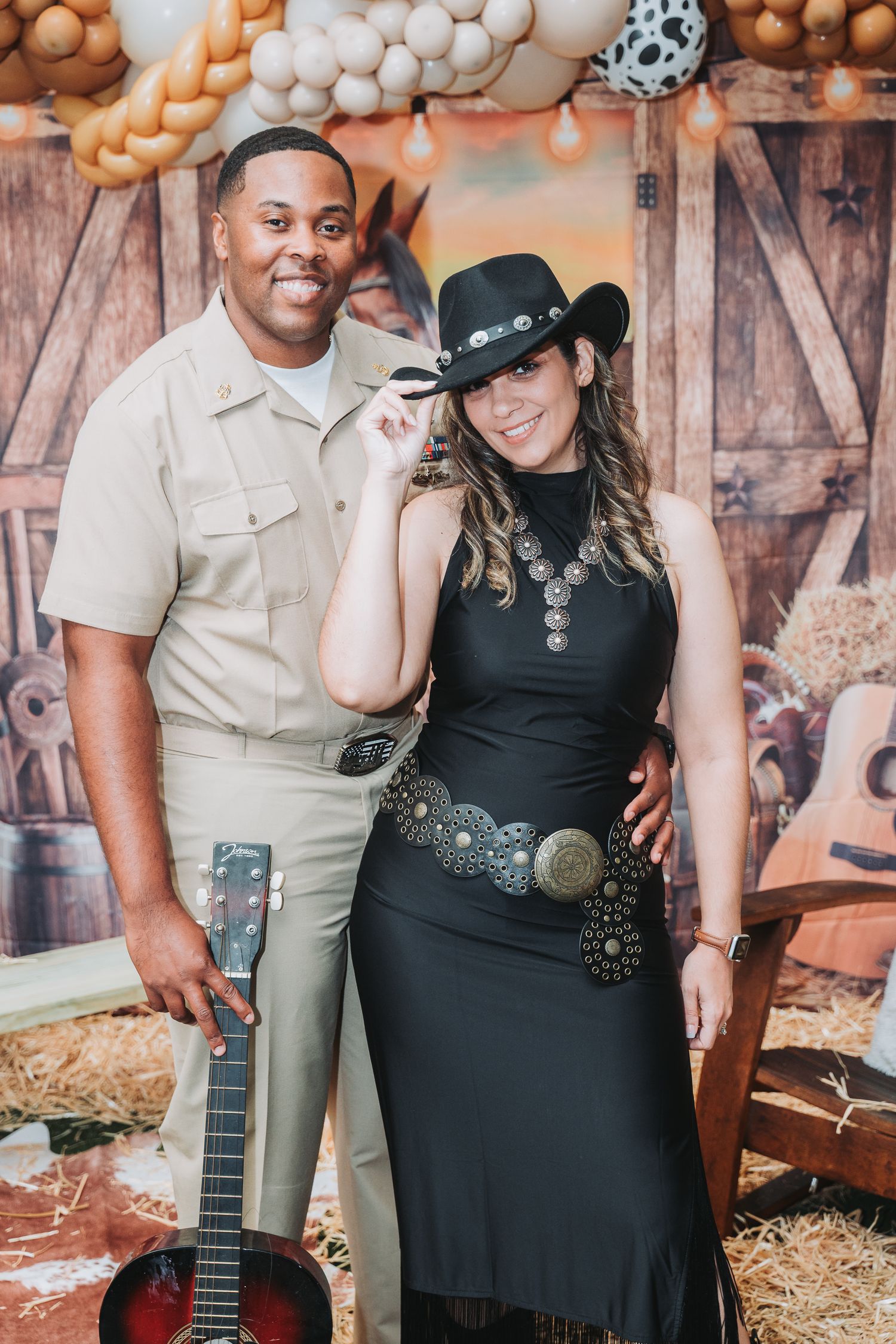 A couple poses in western attire with a guitar against a rustic backdrop with balloon decorations.