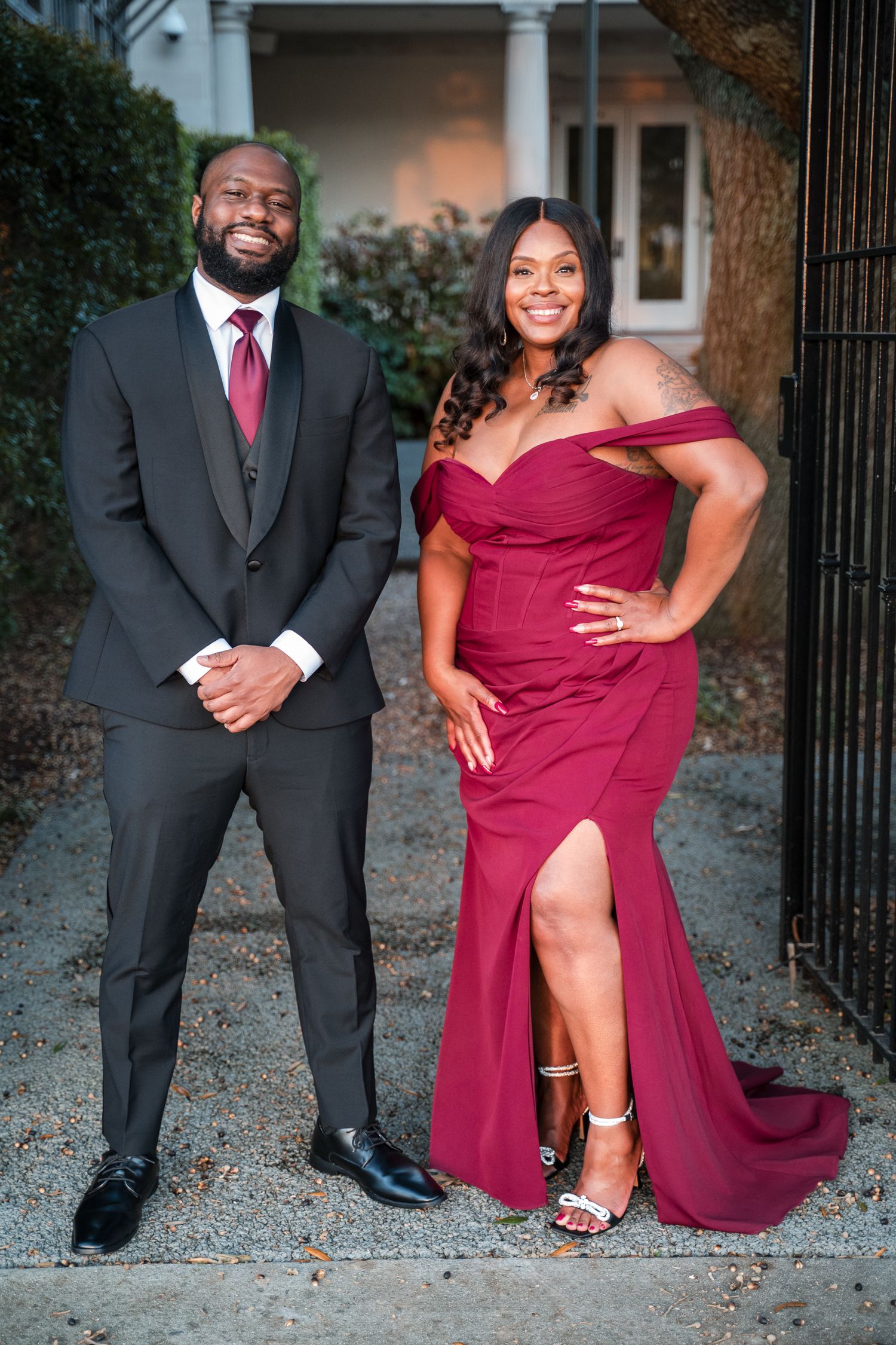 A couple poses together outdoors in formal attire, with a burgundy gown and black suit against a garden backdrop.