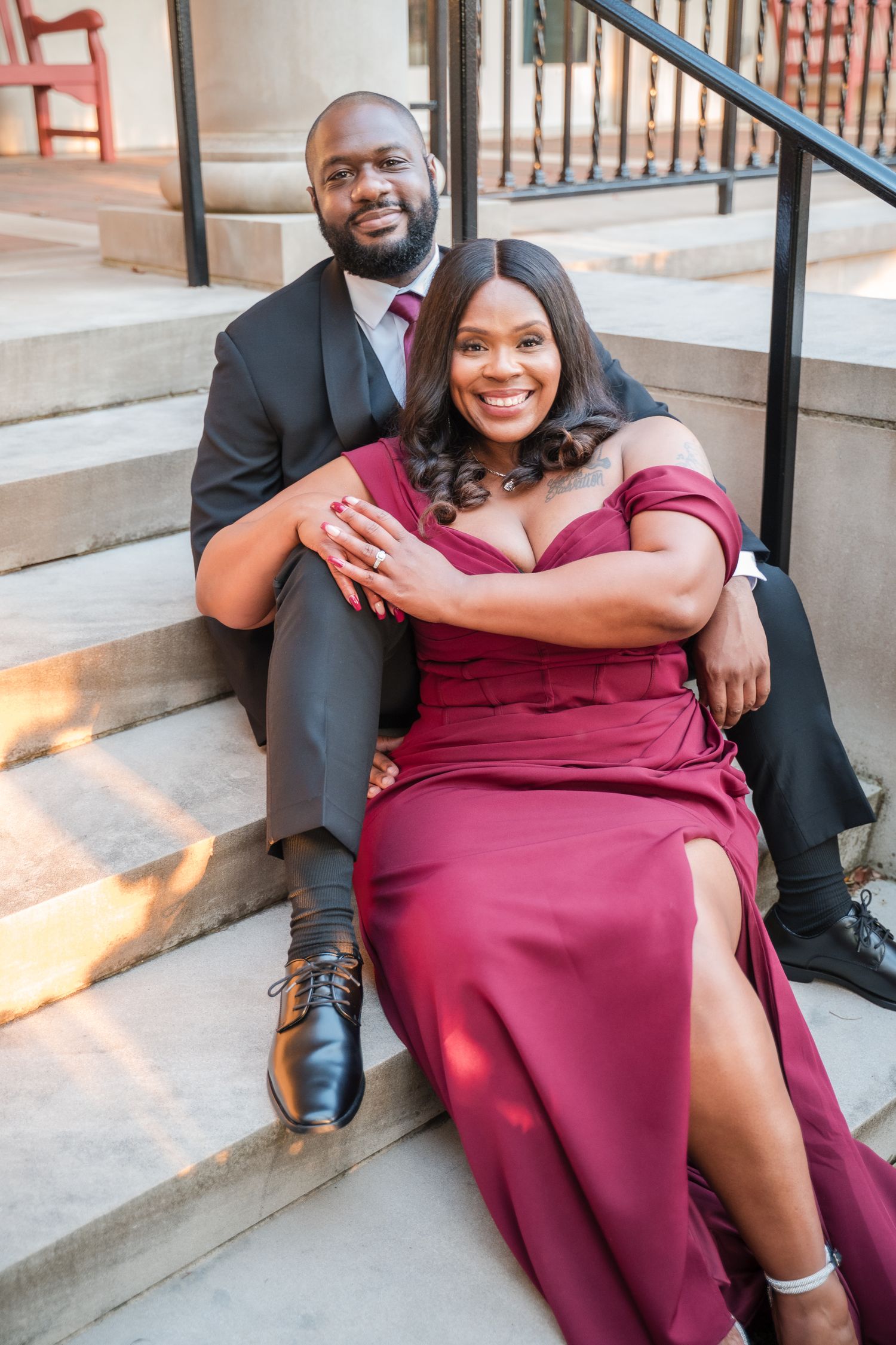 A couple sits together on outdoor steps, with the woman wearing a flowing burgundy dress with a thigh-high slit.