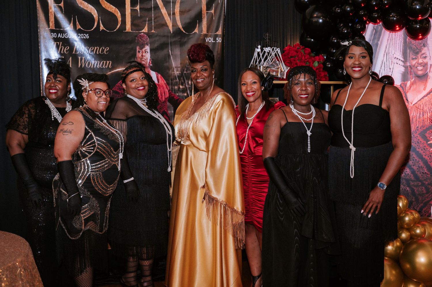 Group of women in elegant black, gold and red evening gowns pose together at a glamorous Essence event with black and gold balloons.