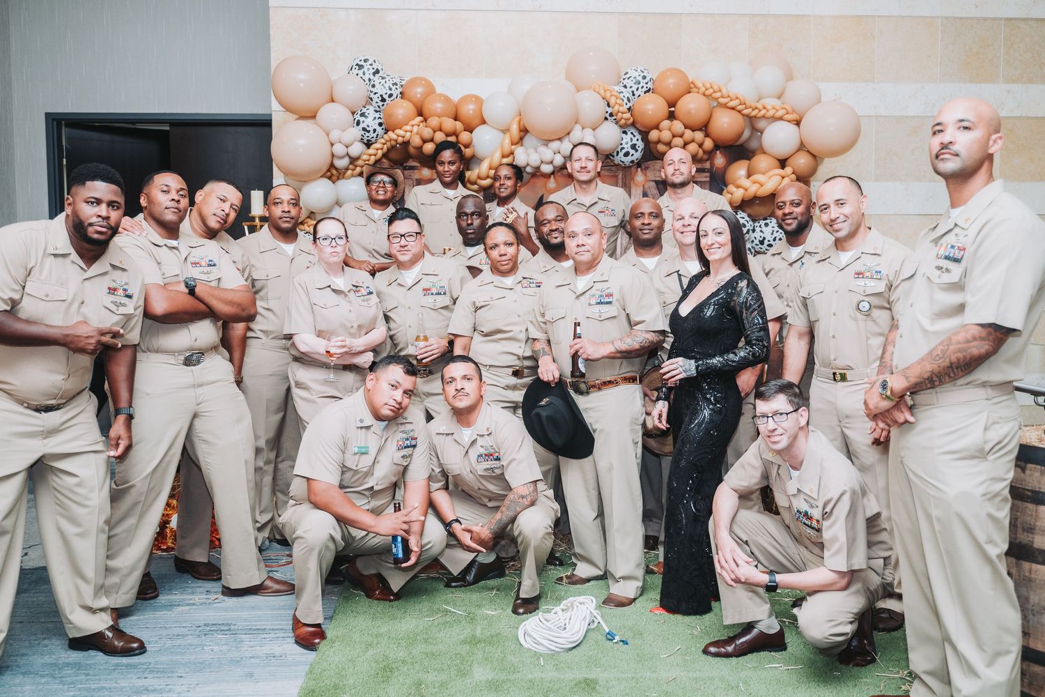 A large group in khaki uniforms poses together in front of a balloon arch at an event.