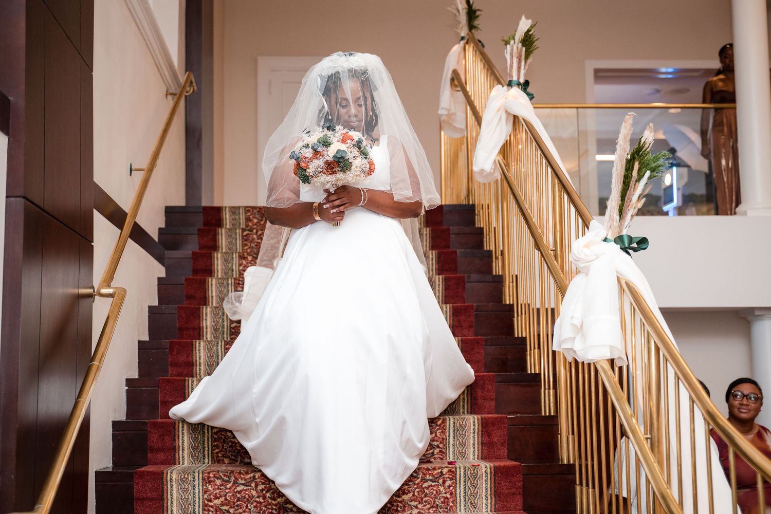 Bride in white wedding gown and veil poses on carpeted staircase holding bouquet.