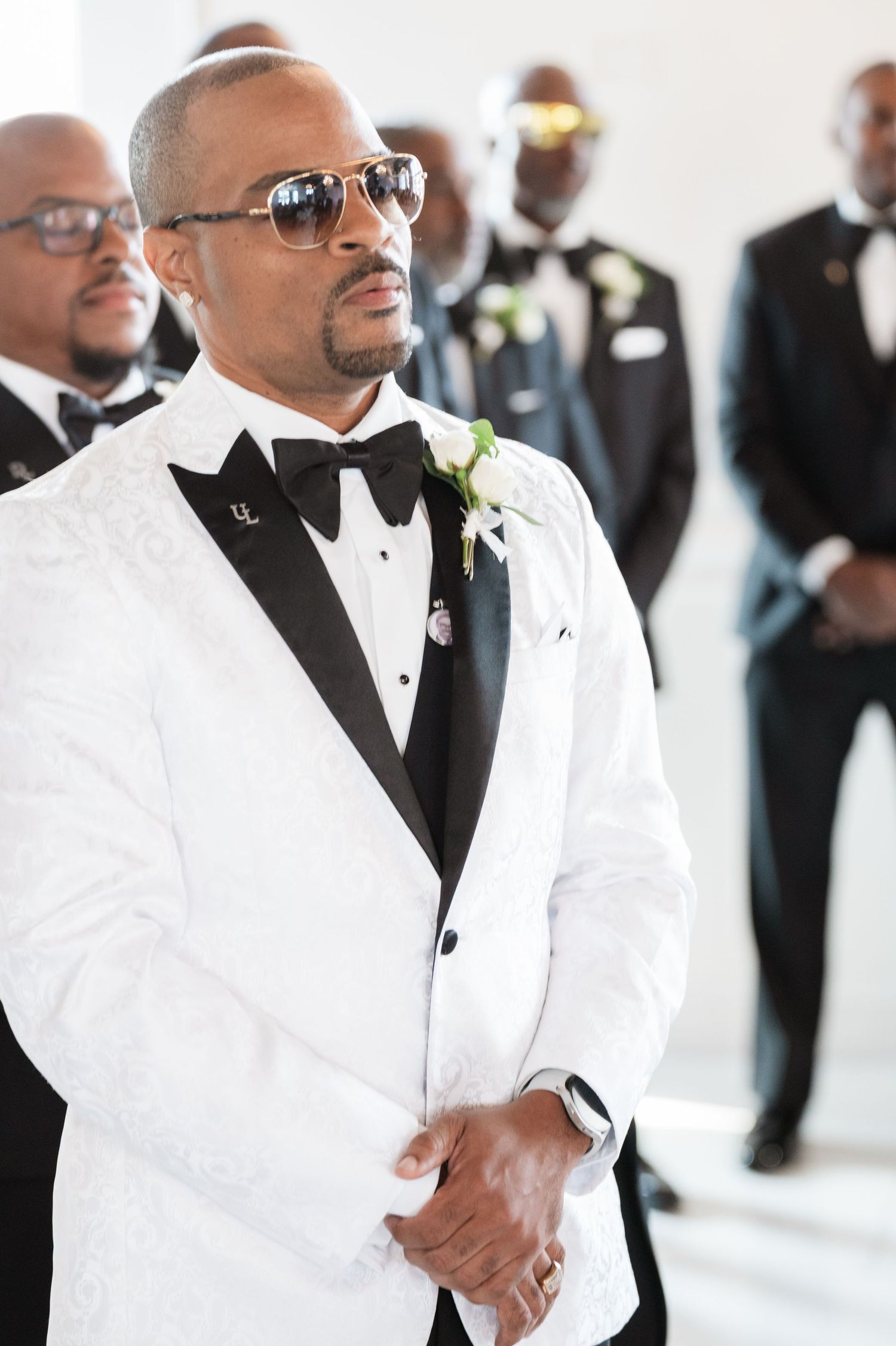 Groomsmen in white tuxedo jackets and black bowties stand together at formal event.