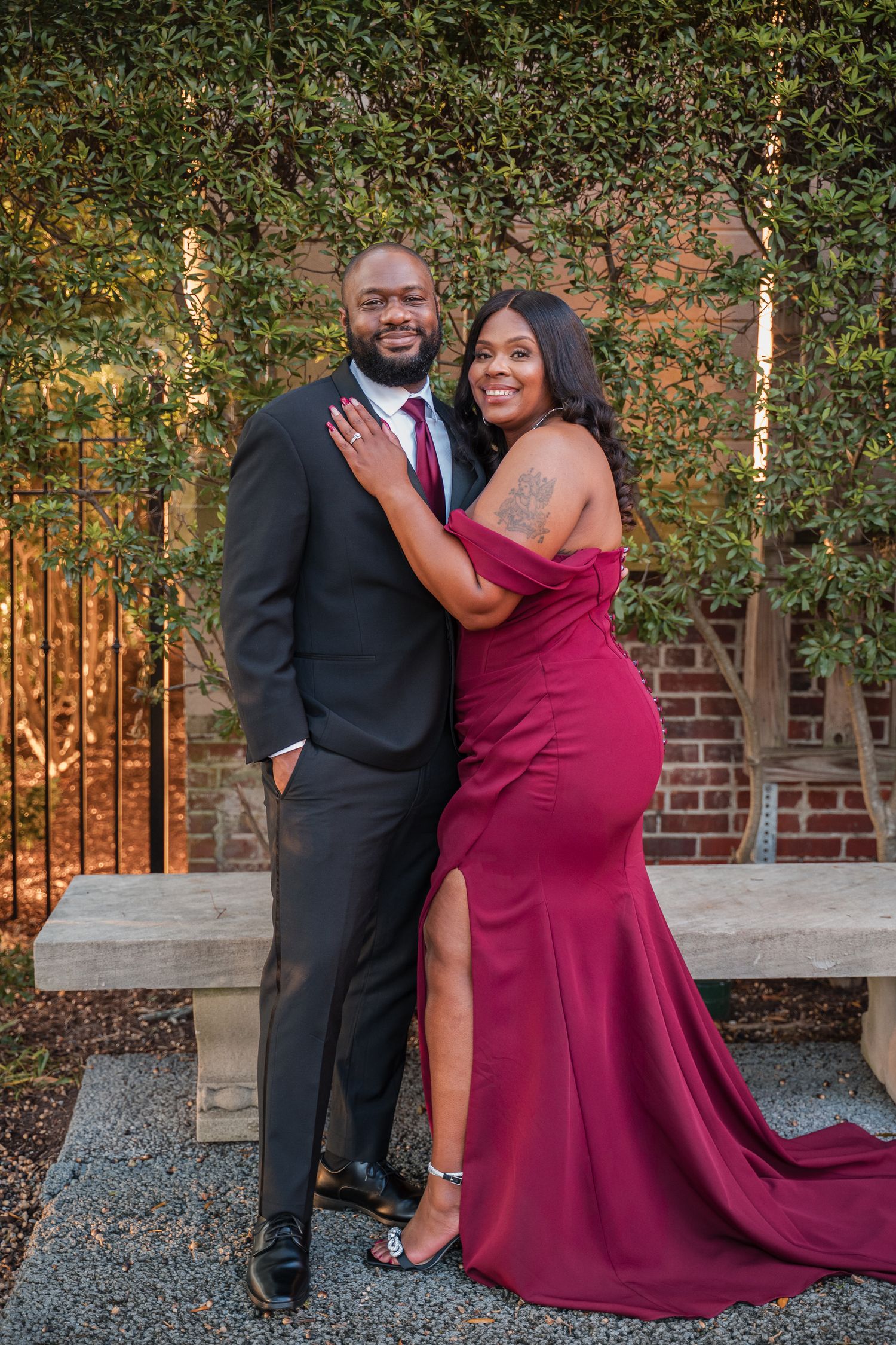 A romantic photoshoot featuring a couple in burgundy and black formal wear against a garden setting.