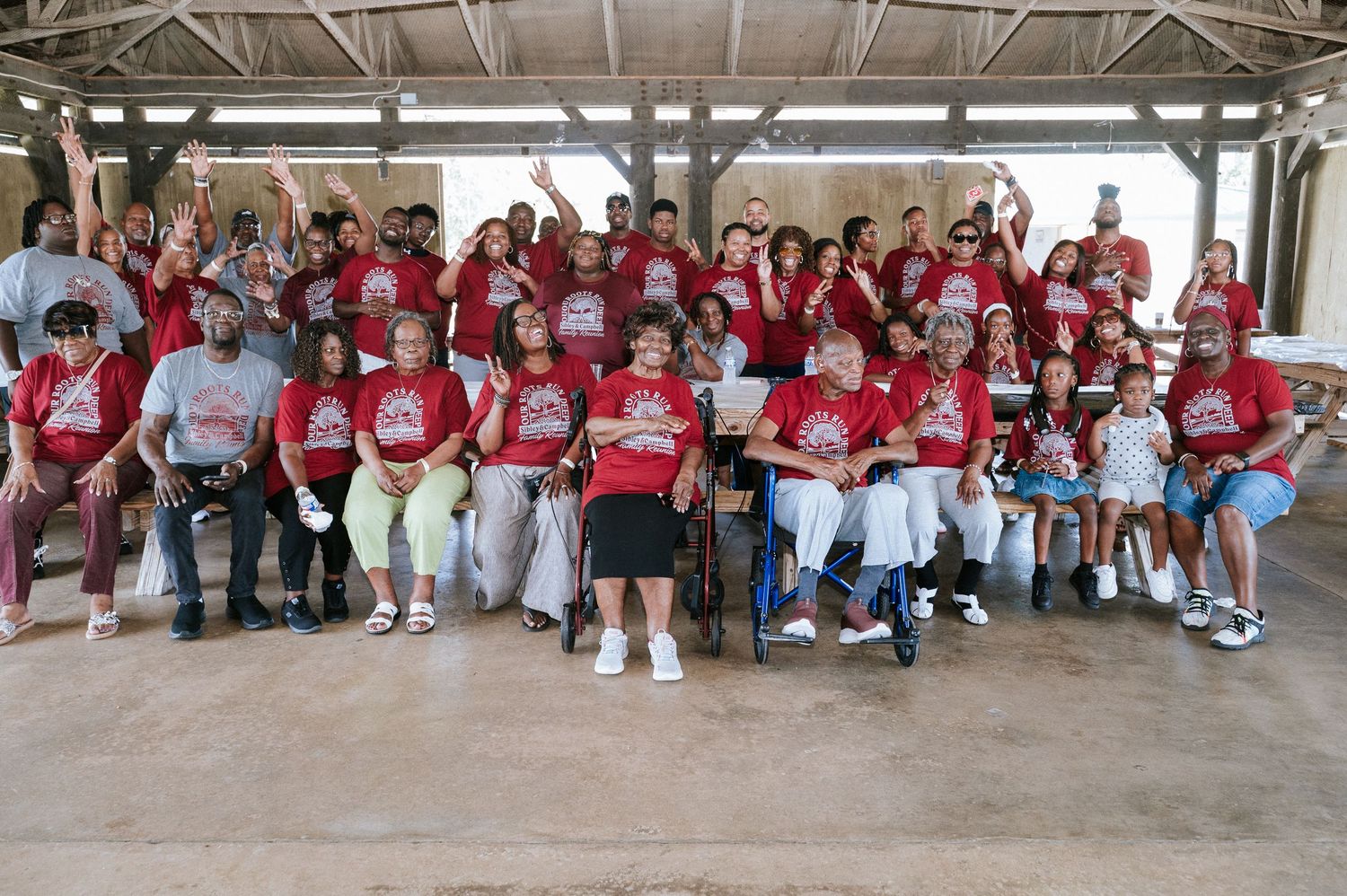 Large group of smiling people in matching red t-shirts posing together under a covered outdoor pavilion, with some seated and others raising