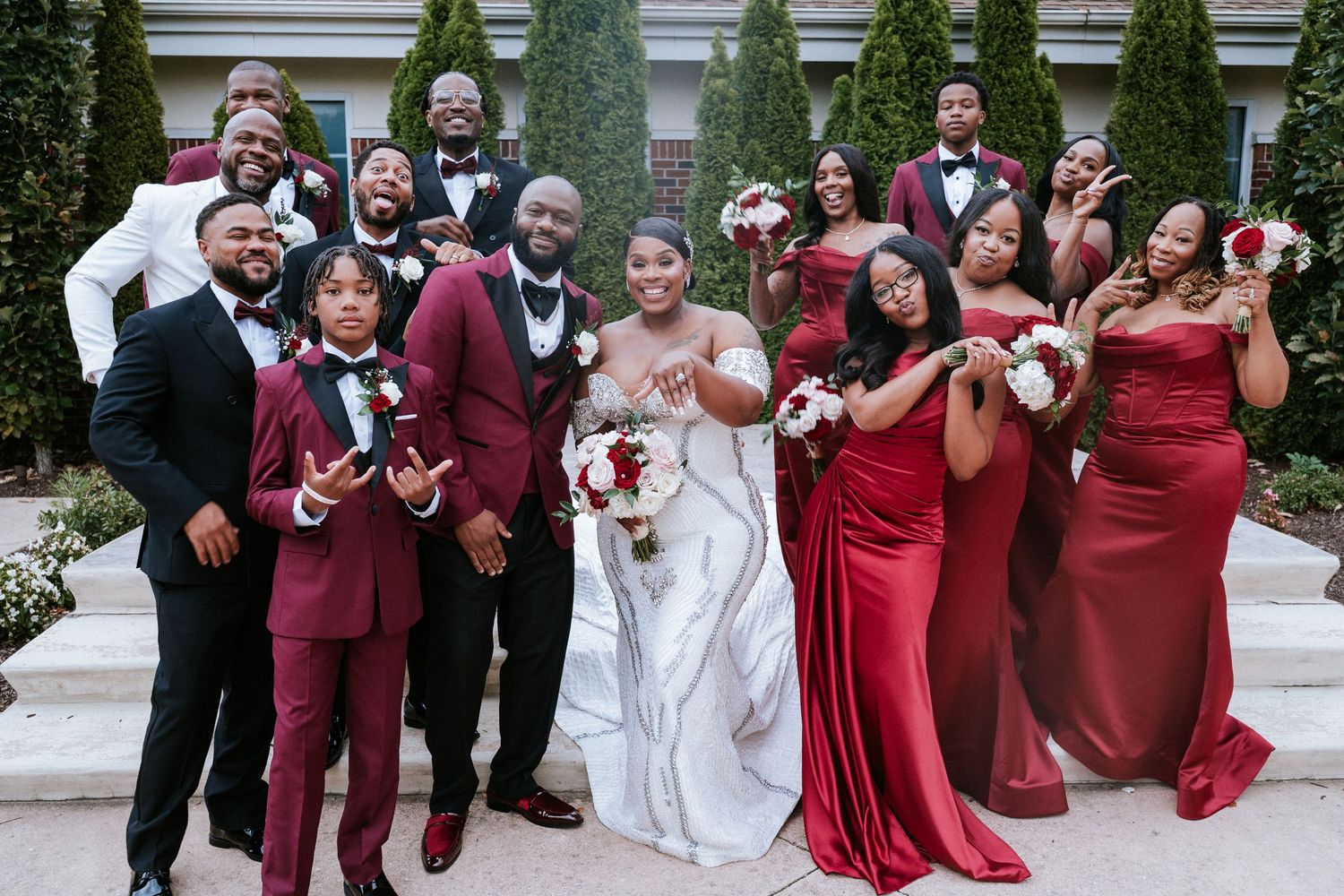 Bride in a white gown holding a bouquet poses with groomsmen in black tuxedos and bridesmaids in burgundy dresses on outdoor steps.