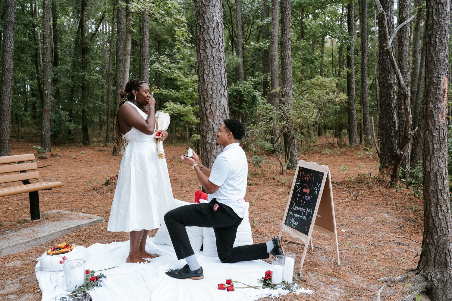 Man in white shirt kneels on one knee holding a ring box while a woman in a white dress covers her mouth in a forest marriage proposal.