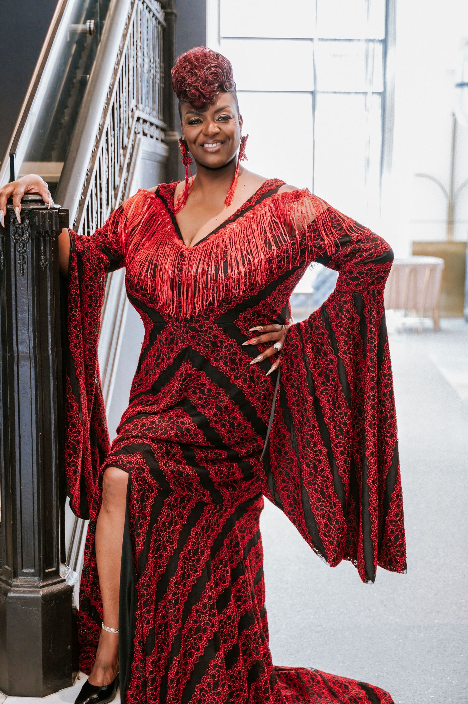 Woman in striking red and black fringe gown with bell sleeves poses gracefully on staircase.