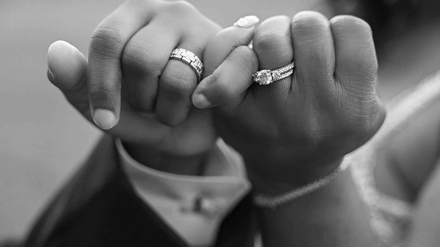 Close-up black and white photo of two hands with engagement rings making pinky promise gesture.